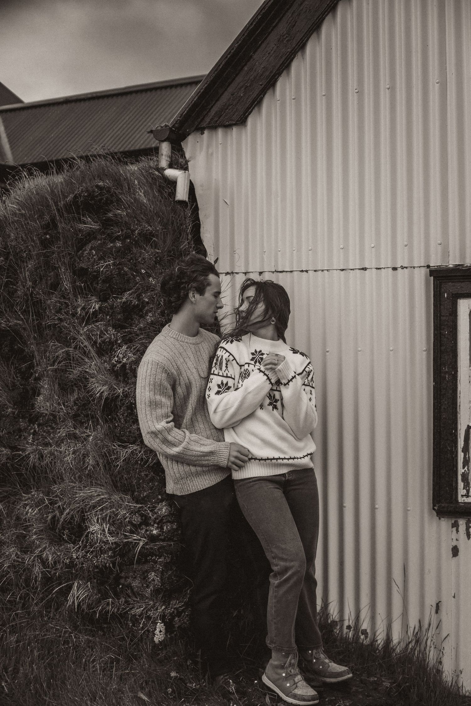 Black and white photo of a couple in sweaters embracing against a corrugated metal wall.