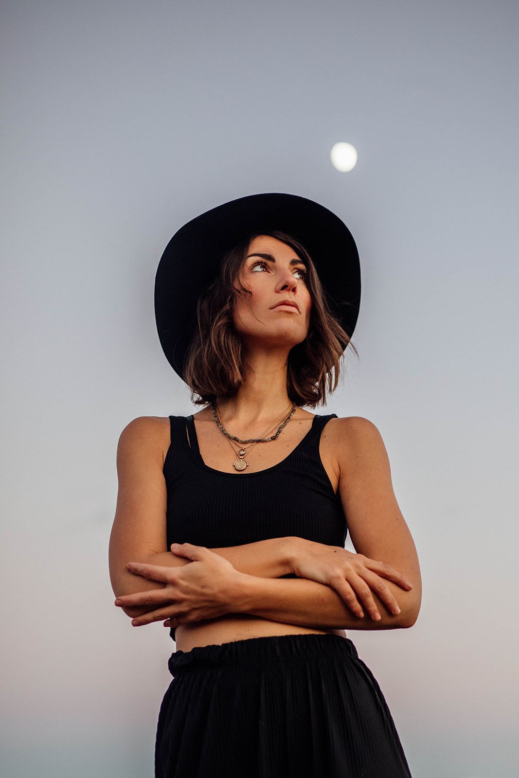 Artistic portrait of someone in black attire and hat gazing up at the moon against a dusky sky.