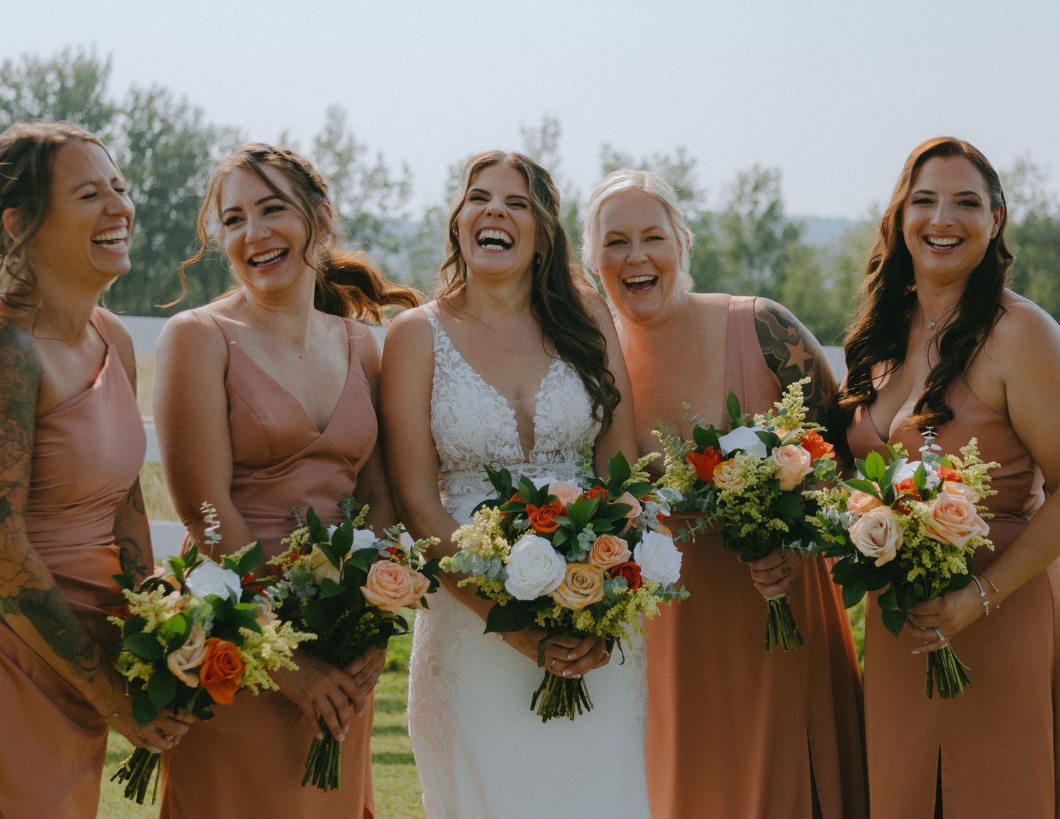 A wedding party with bridesmaids in peach-colored dresses holding vibrant bouquets of orange and yellow flowers sharing joyful laughter.