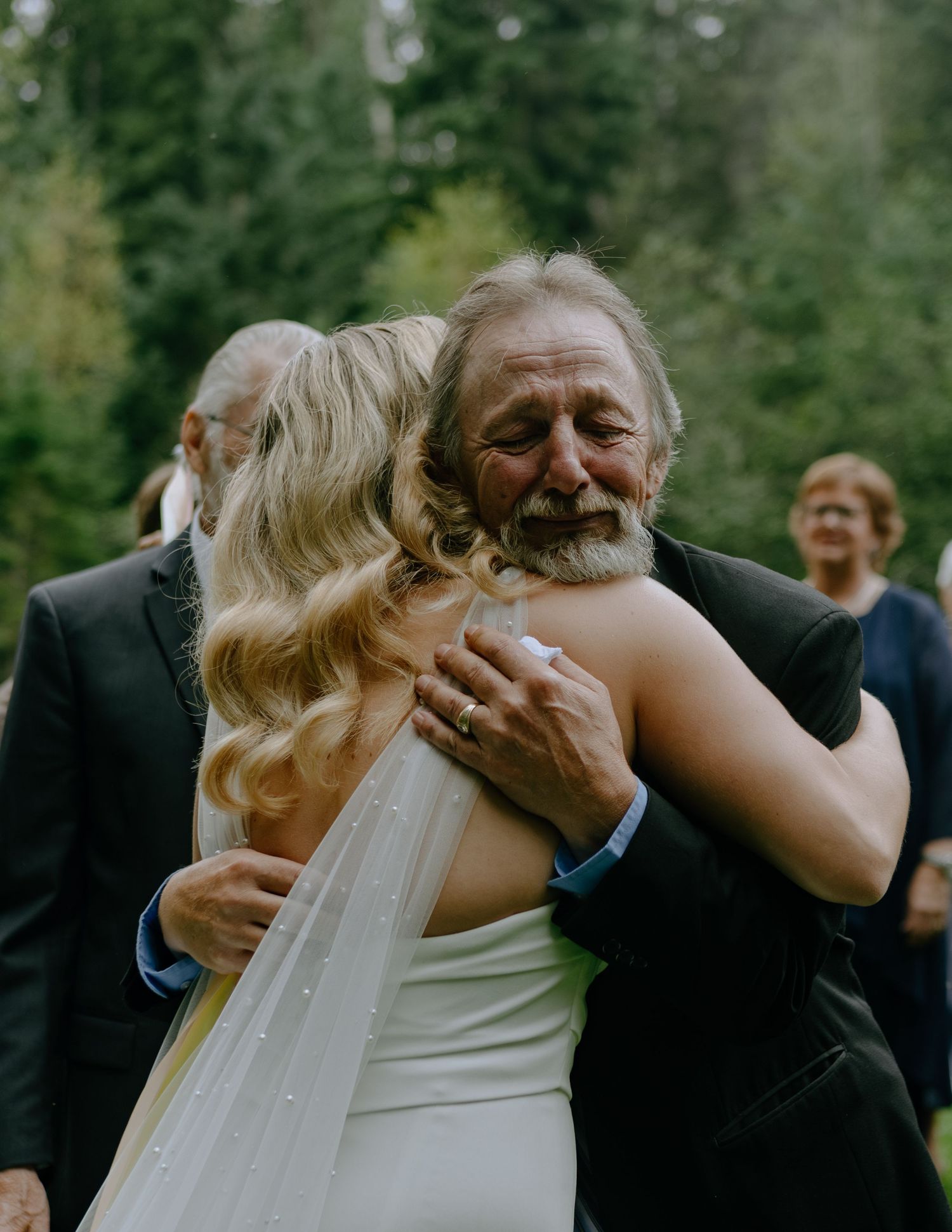 An emotional embrace between two people at an outdoor wedding ceremony with trees in the background.