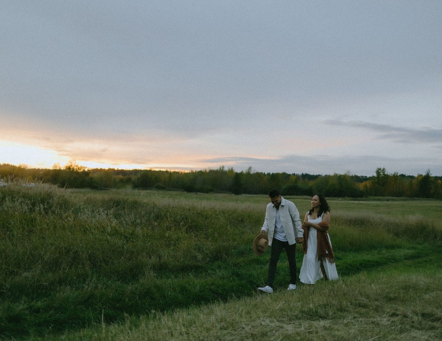 Couple walking hand in hand through a meadow at sunset with moody sky overhead.