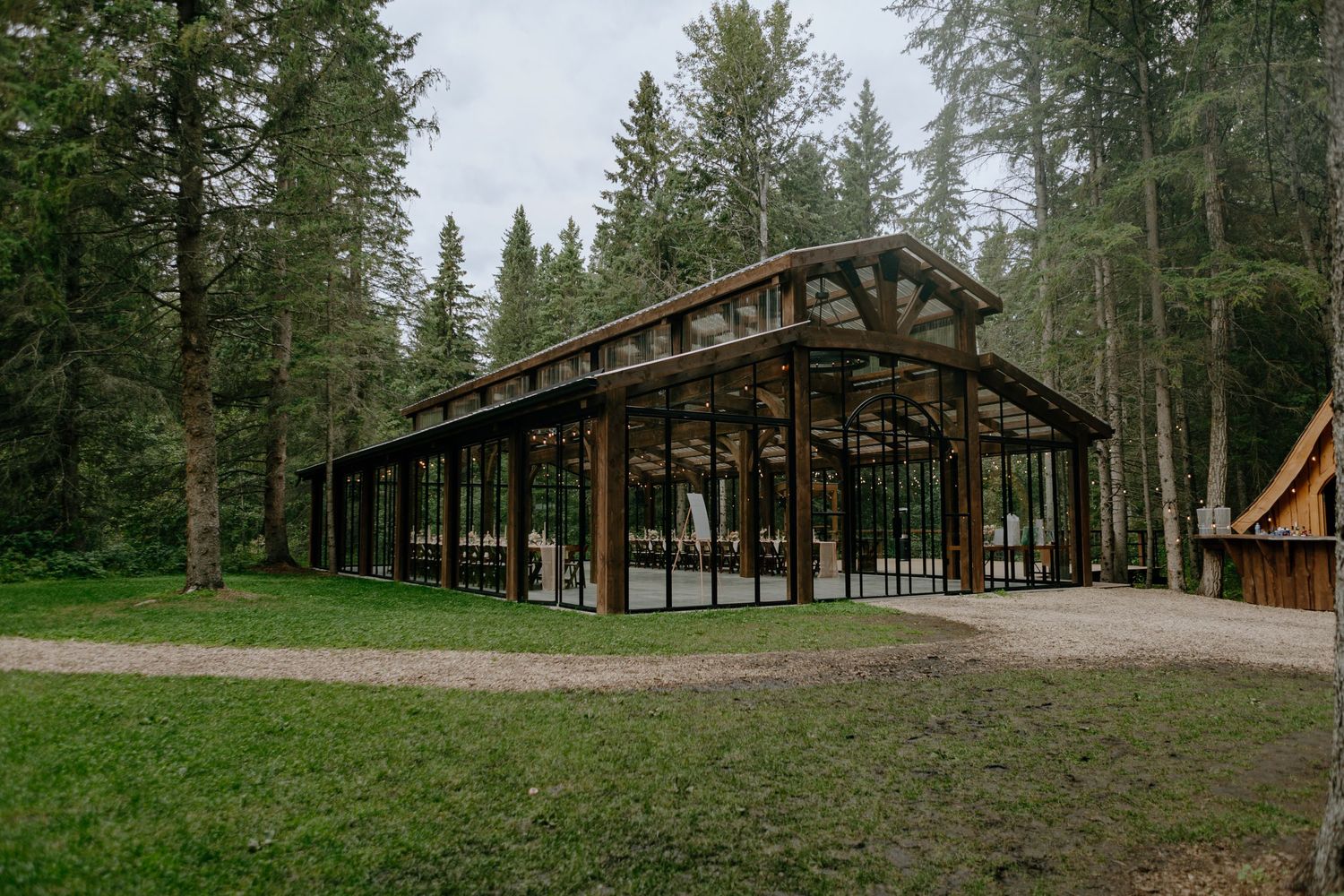 A modern glass-walled pavilion structure with exposed wooden beams sits nestled among tall evergreen trees in a forest setting.