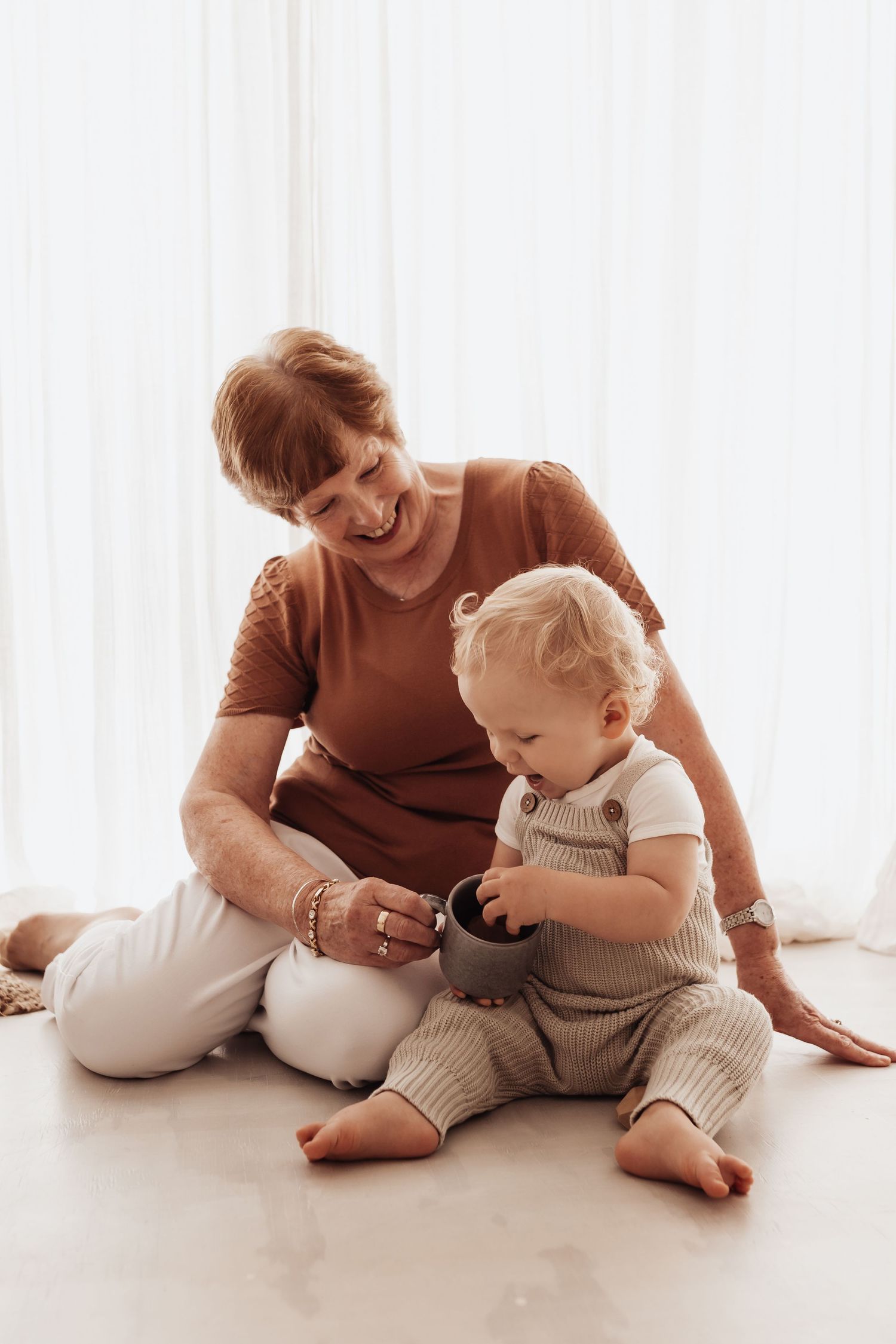 Adult in brown sweater sitting on floor playing with toddler wearing gray overalls in bright minimal room setting.