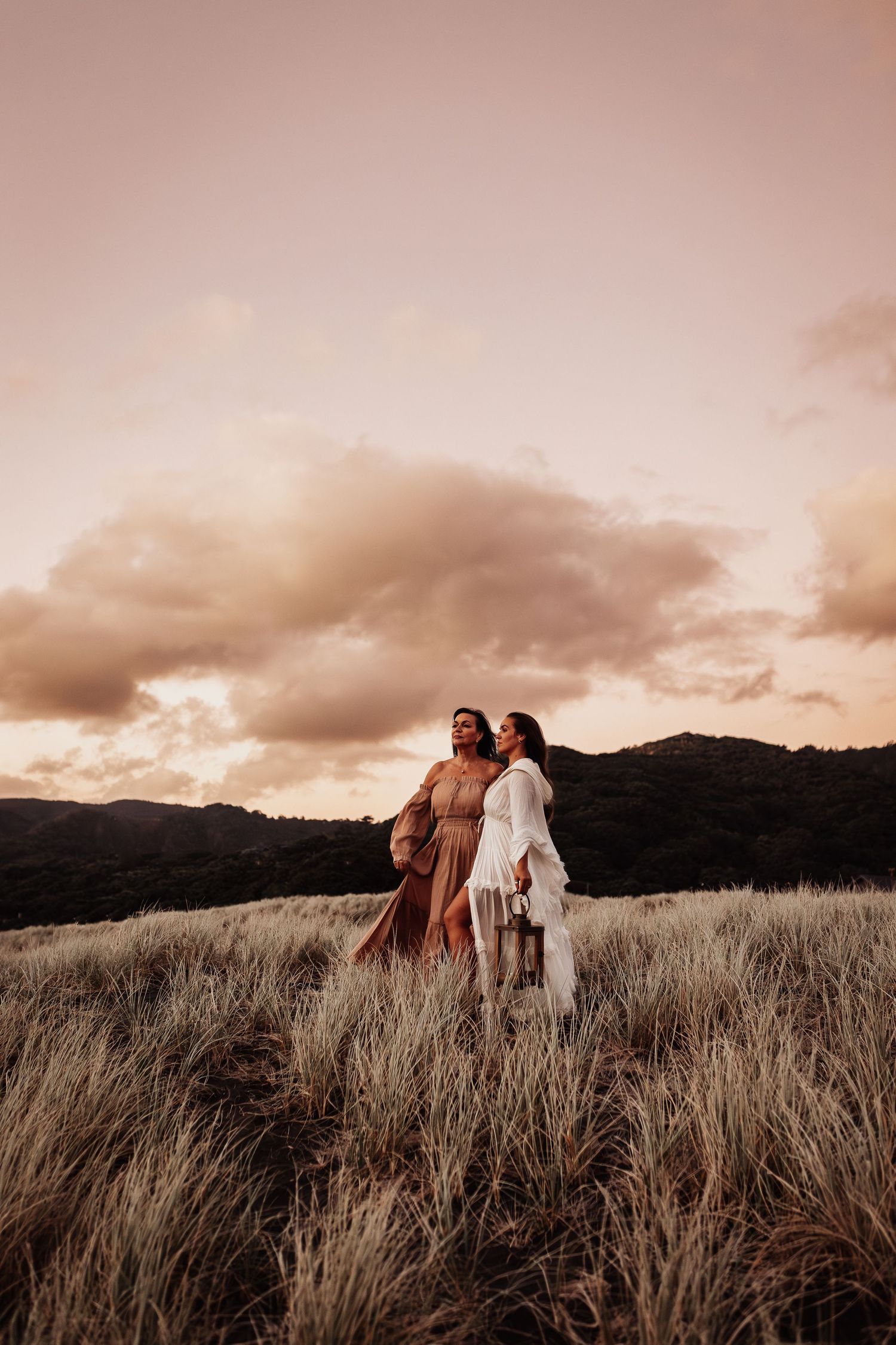 Two people in flowing dresses stand together in tall grass field at sunset with mountains in background.