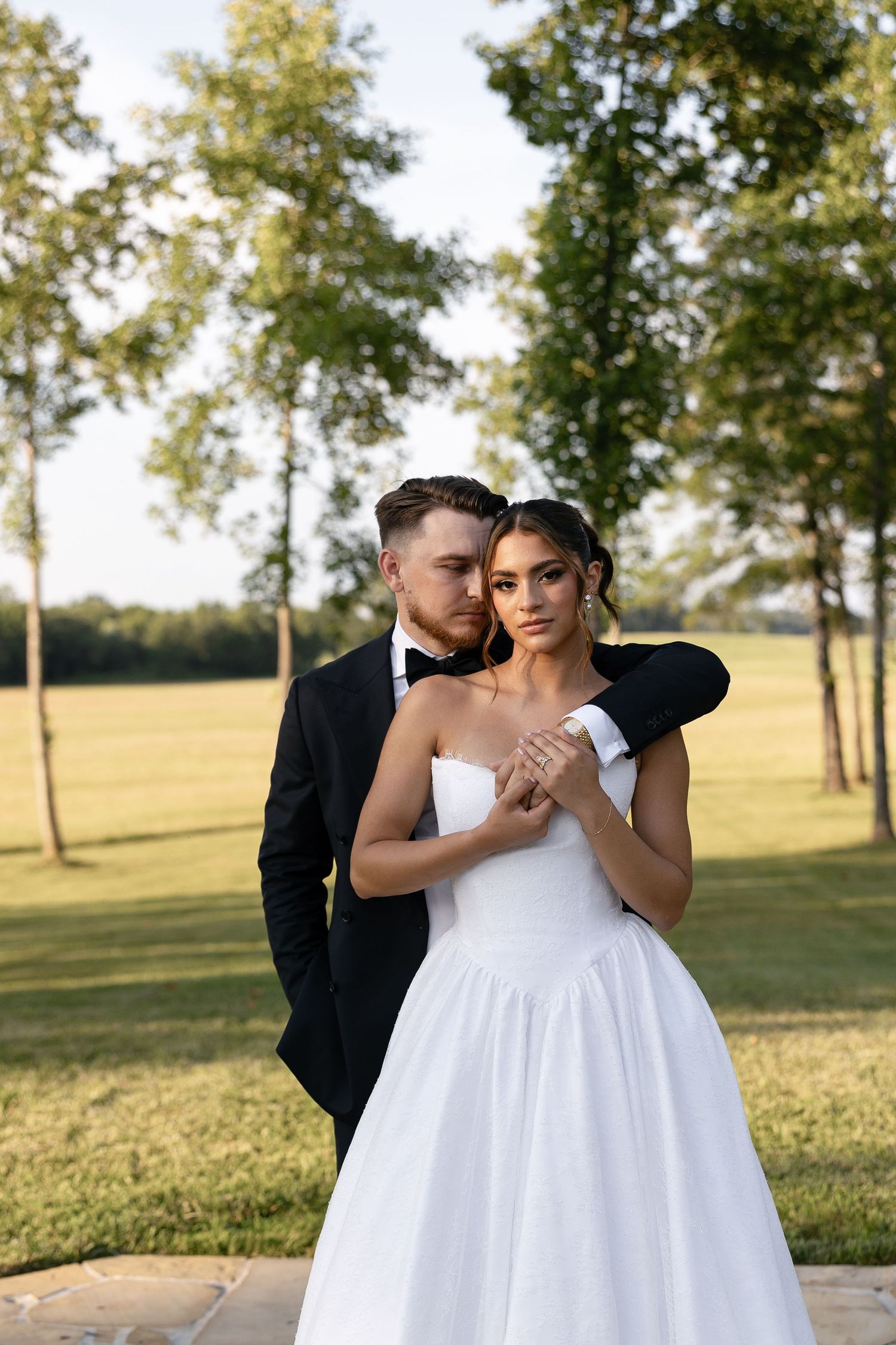 A couple embraces outdoors in romantic wedding attire on a sunny day with trees in the background.