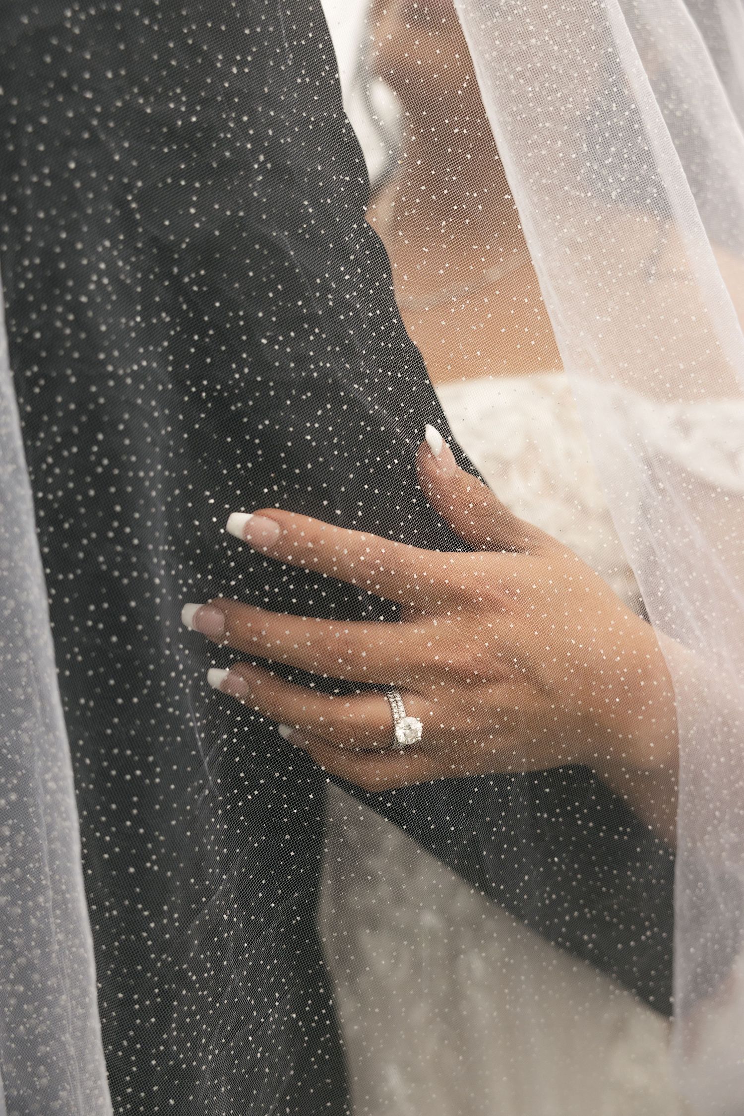 A hand wearing an engagement ring delicately touches a sheer white veil against a dark background with sparkles.