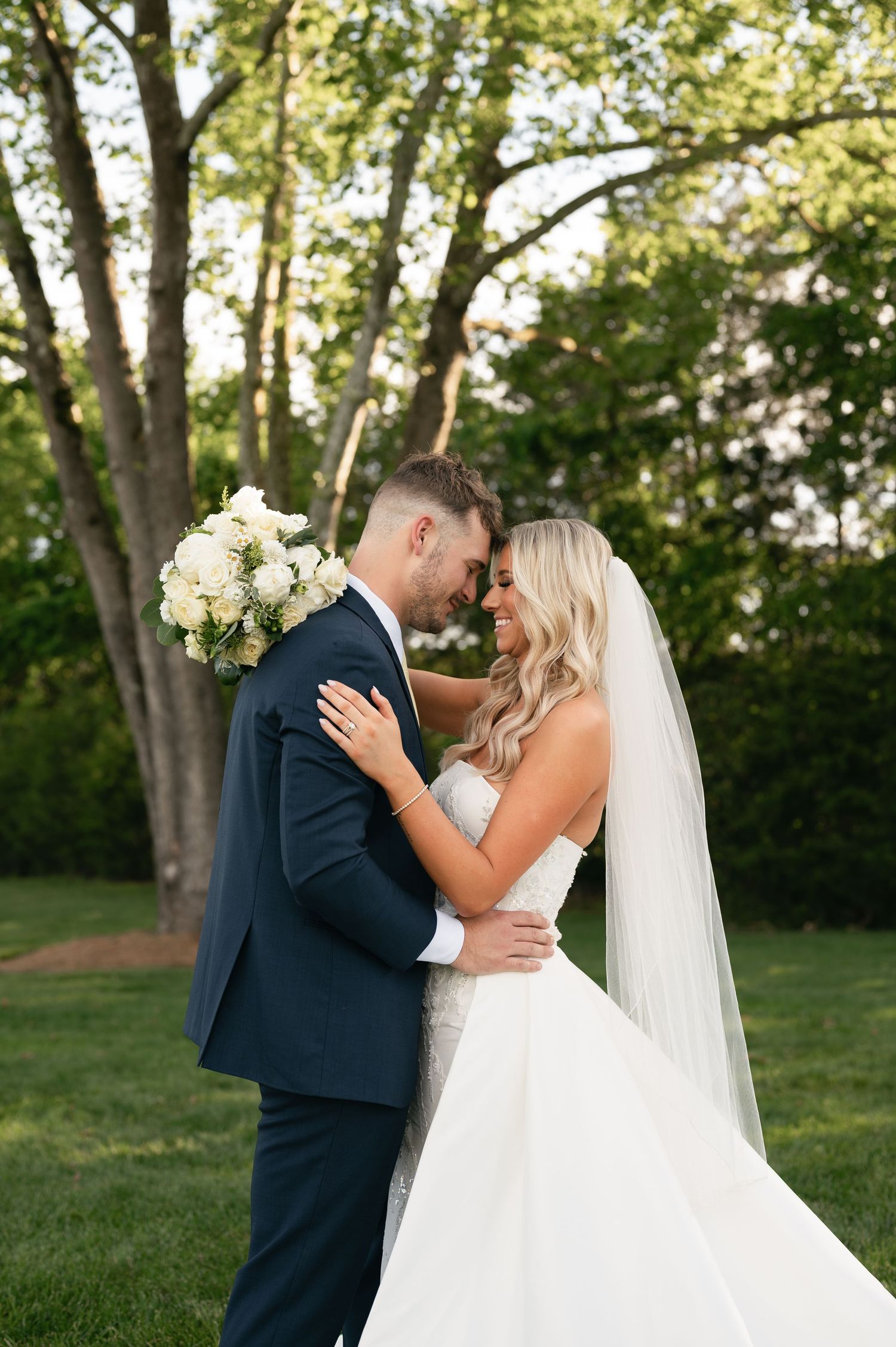 Bride in flowing white gown and groom in navy suit embrace outdoors under sunlit trees with white rose bouquet.