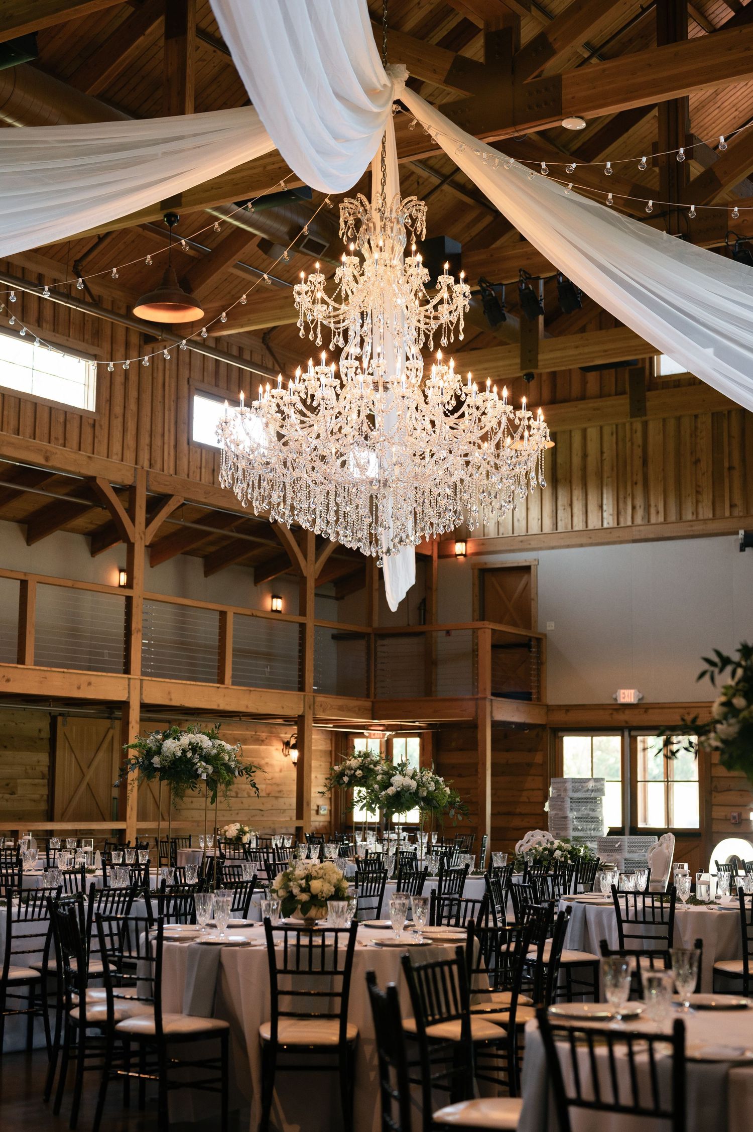 Elegant barn wedding reception venue with crystal chandelier, white draping, and round tables set for dining.
