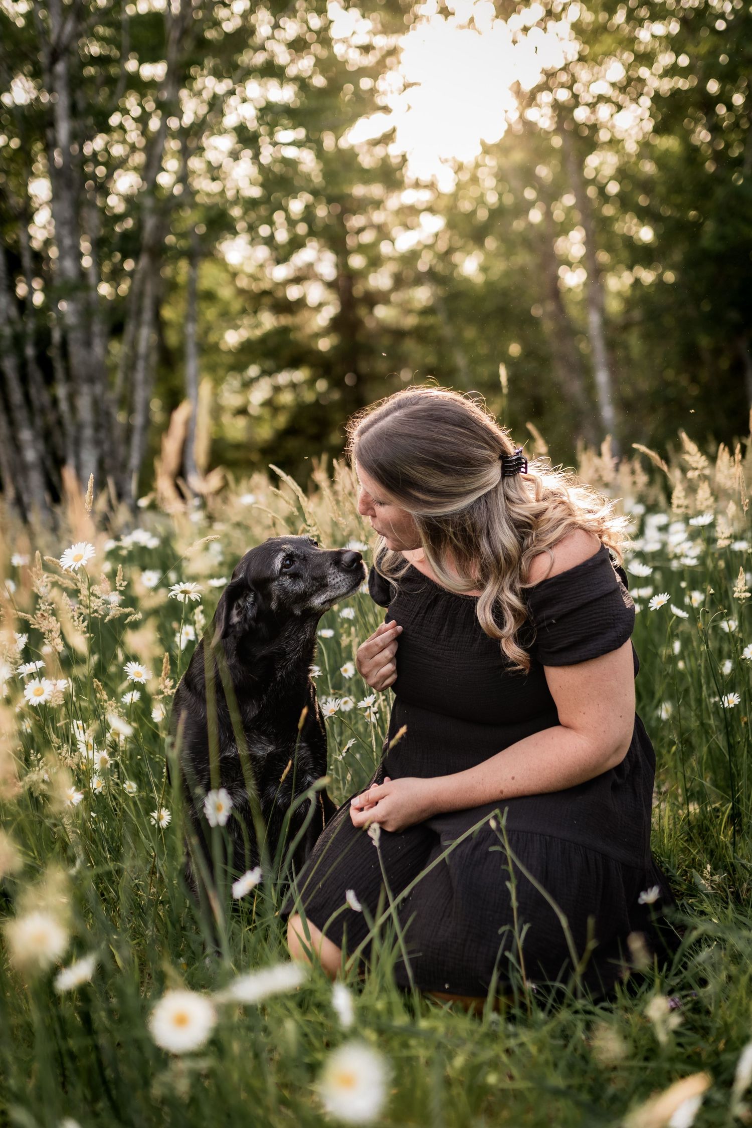 Woman in black dress sharing tender moment with dog among blooming daisies at sunset.