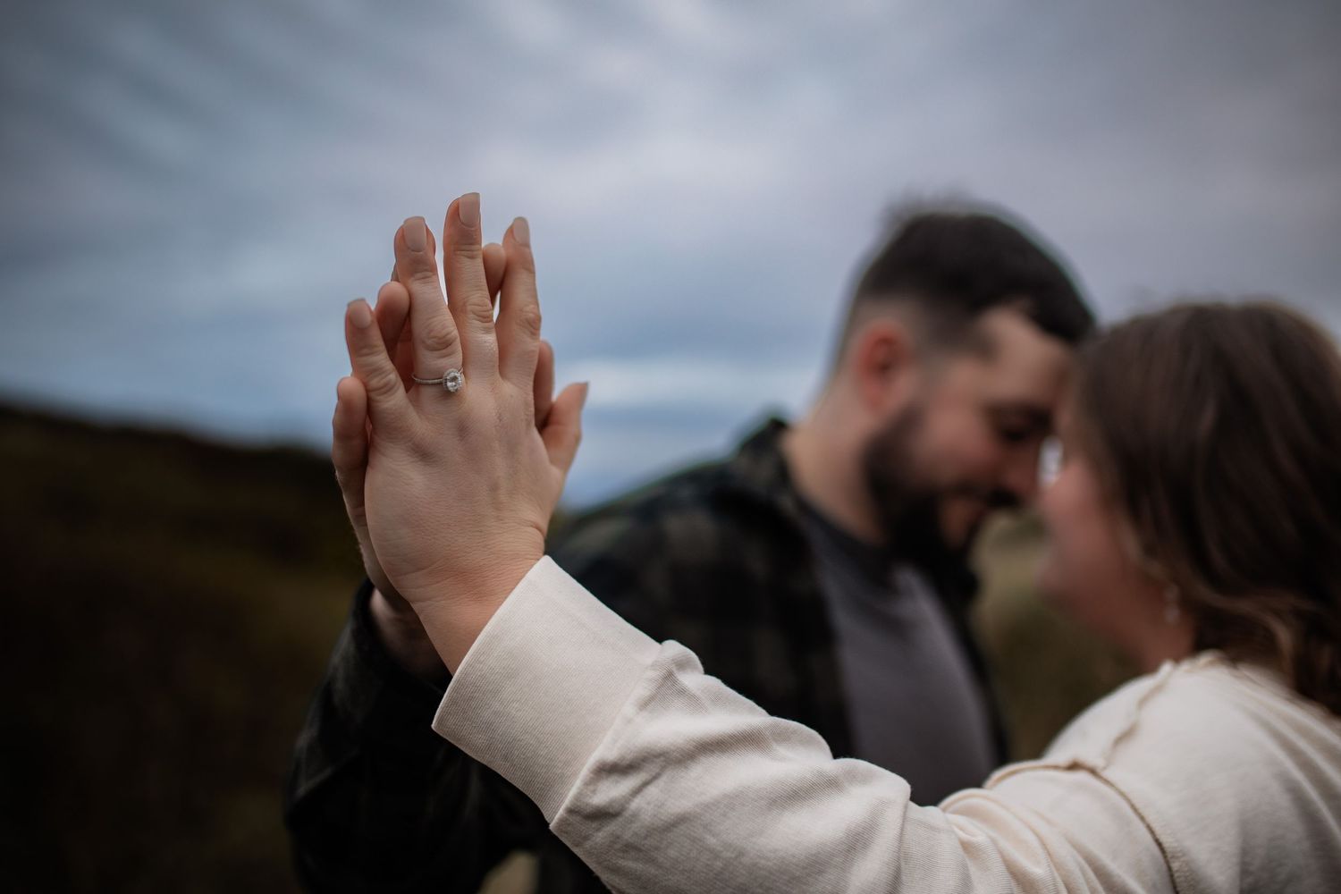 A couple holds hands and touches palms during an intimate moment outdoors beneath a moody sky.