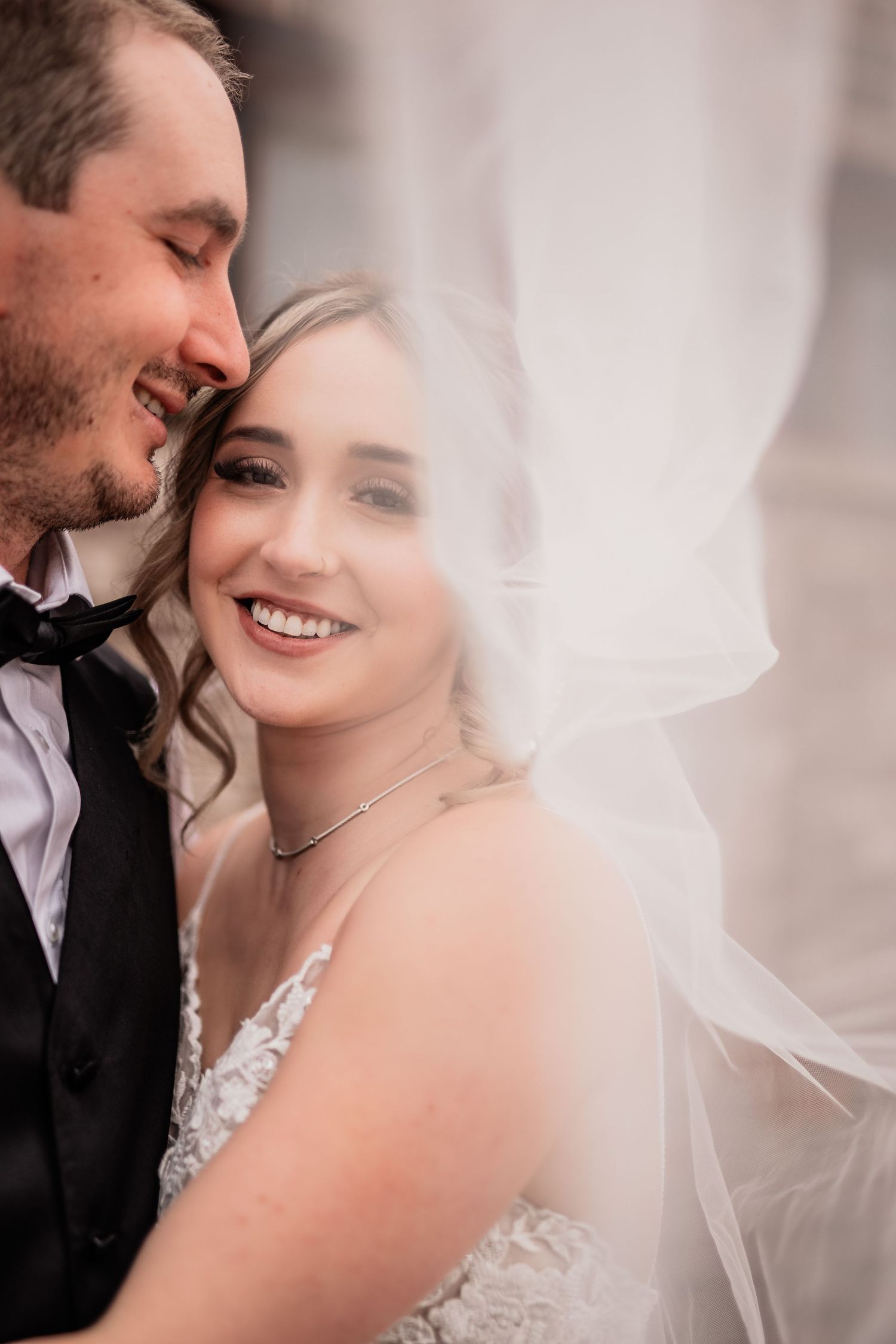 Newlywed couple shares intimate moment with bride in lace dress and flowing veil during wedding photoshoot.
