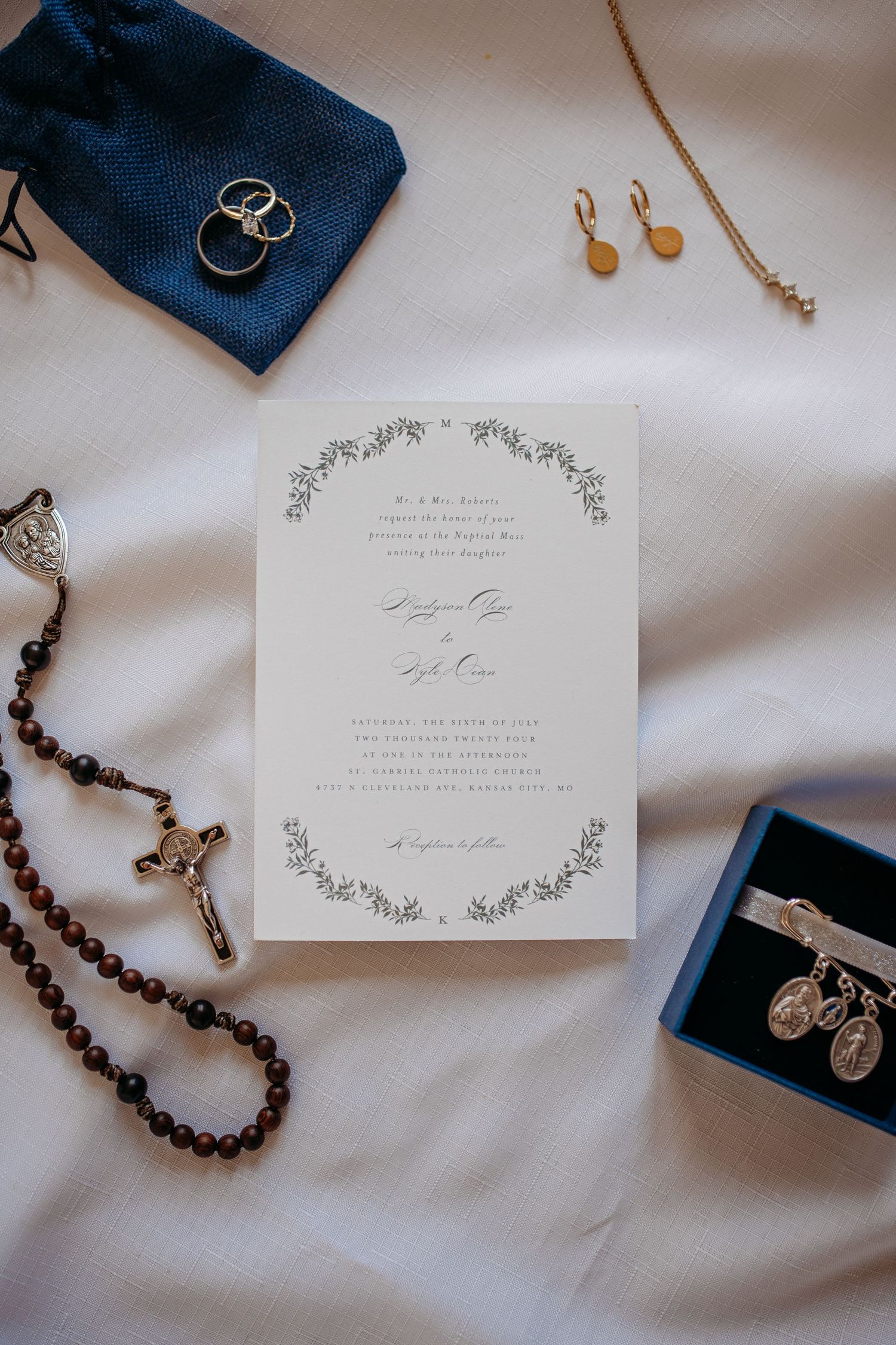 Detail shot of wedding invitation displayed with jewelry and Catholic rosary beads on white fabric, featuring saint medals in St. Louis, MO.