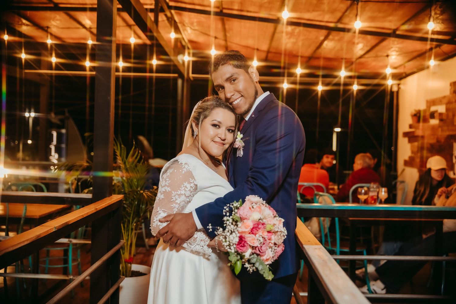 Couple embracing in romantic evening setting with string lights and floral bouquet.