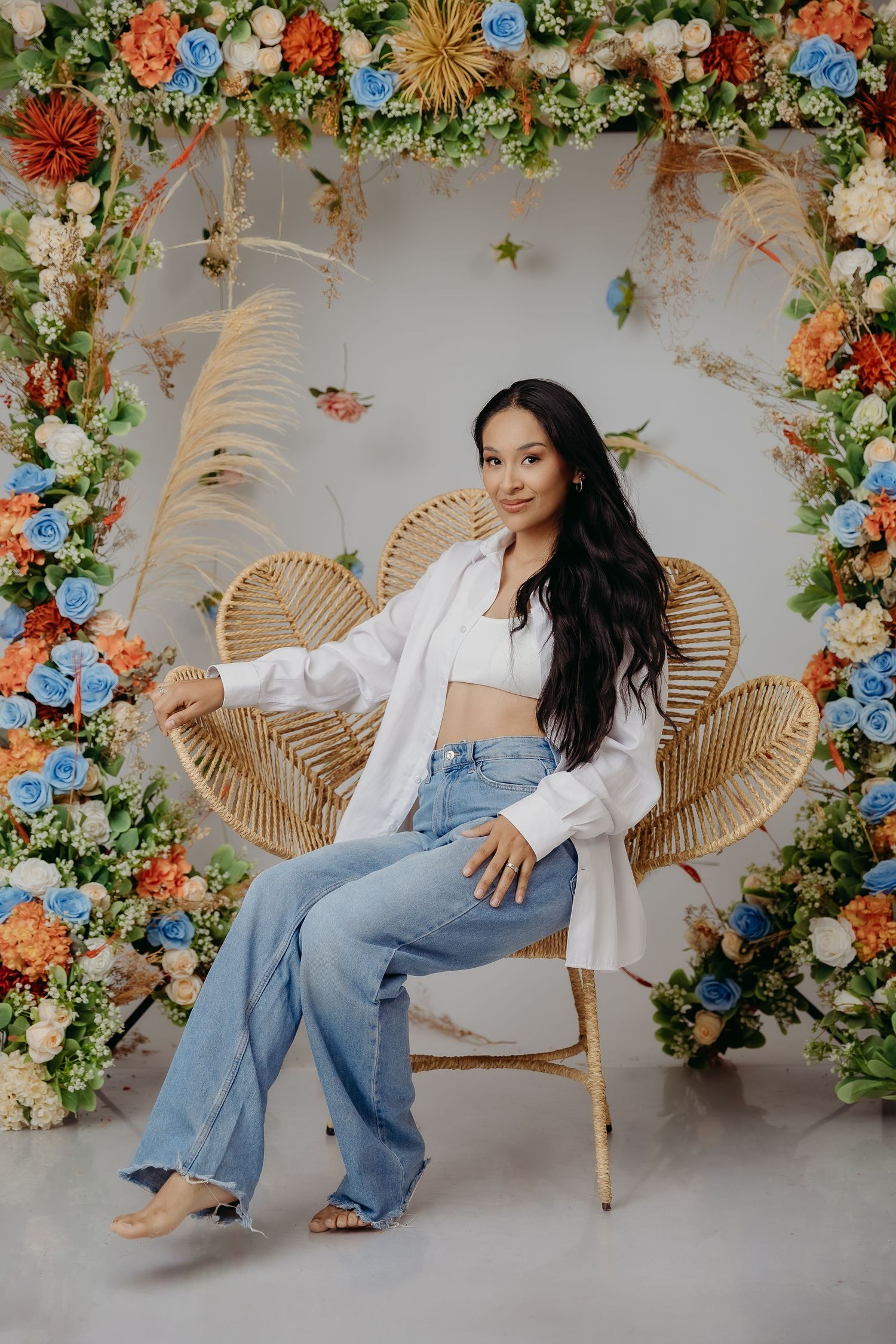 Rattan peacock chair surrounded by floral arch with blue and orange blooms creates dreamy boho photo backdrop.