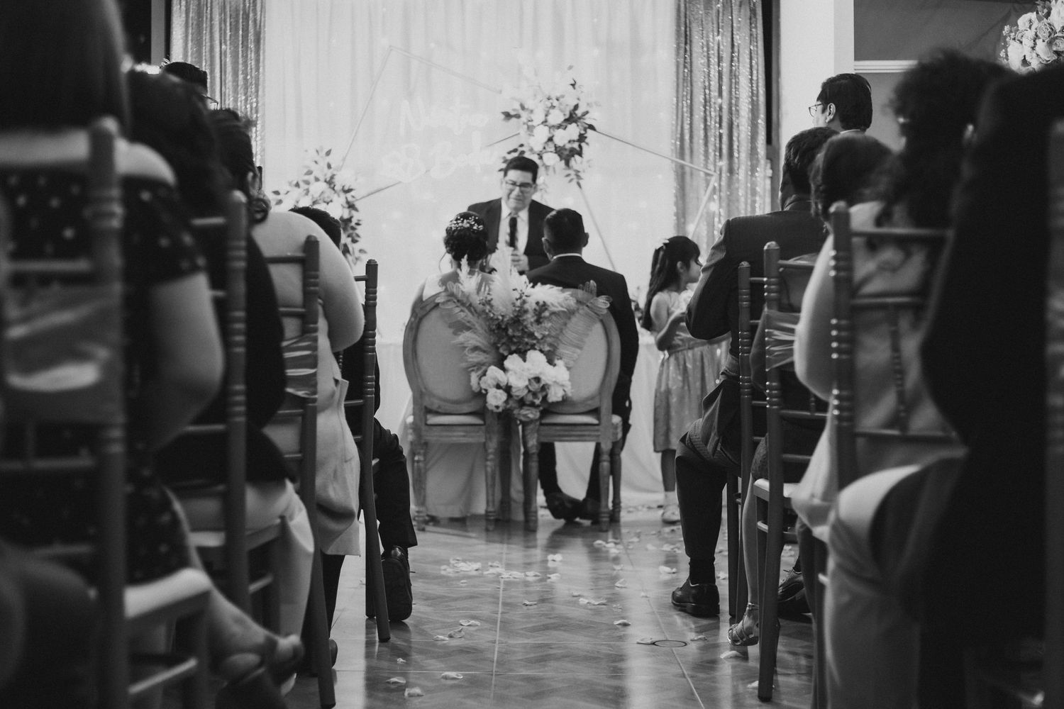 Black and white photo of indoor wedding ceremony with guests seated in rows and floral decorations.