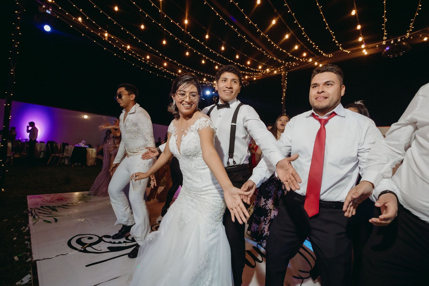 Wedding party dancing on illuminated dance floor under string lights at outdoor reception.