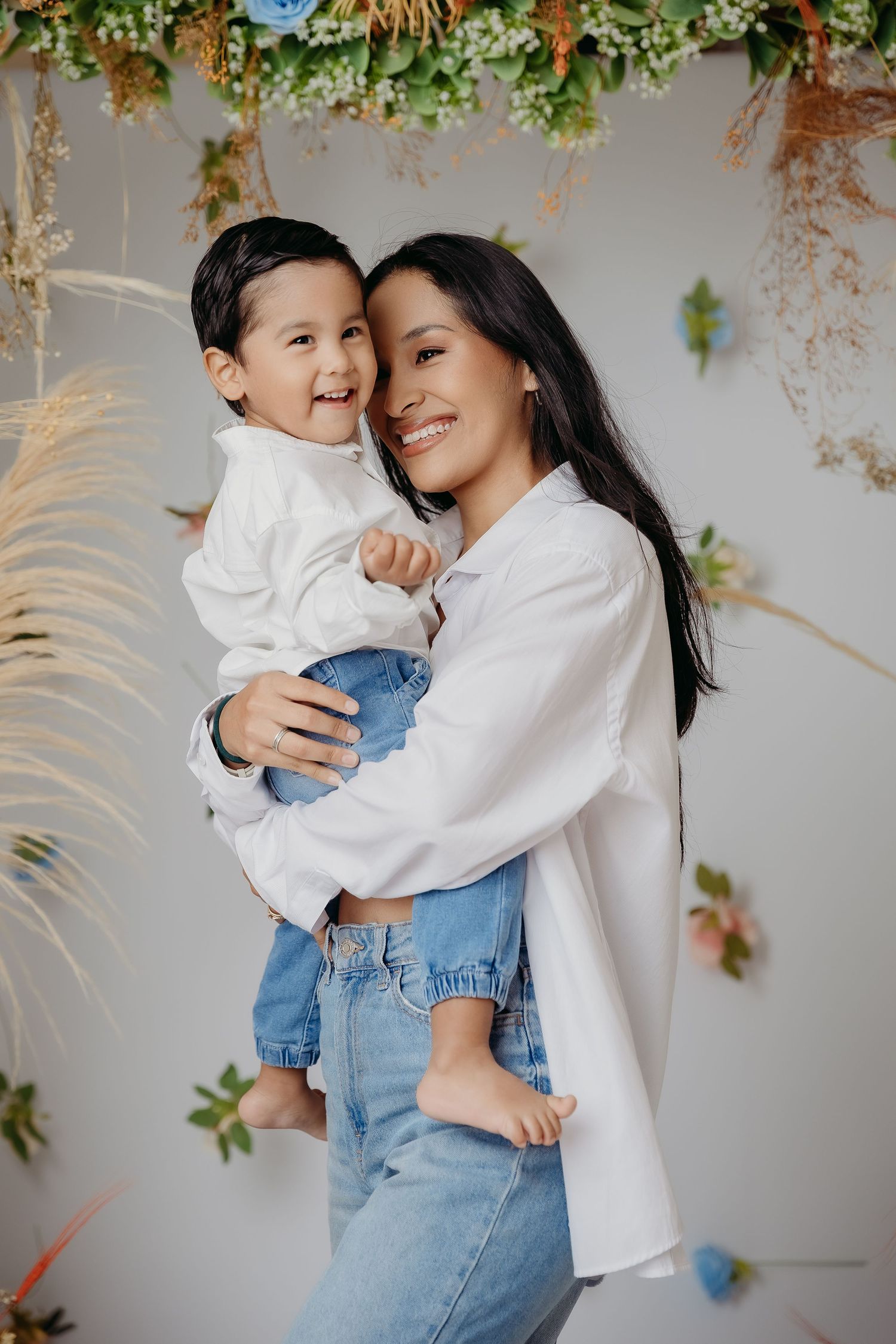 Portrait of mother and child in white tops and denim embracing and laughing against floral backdrop.