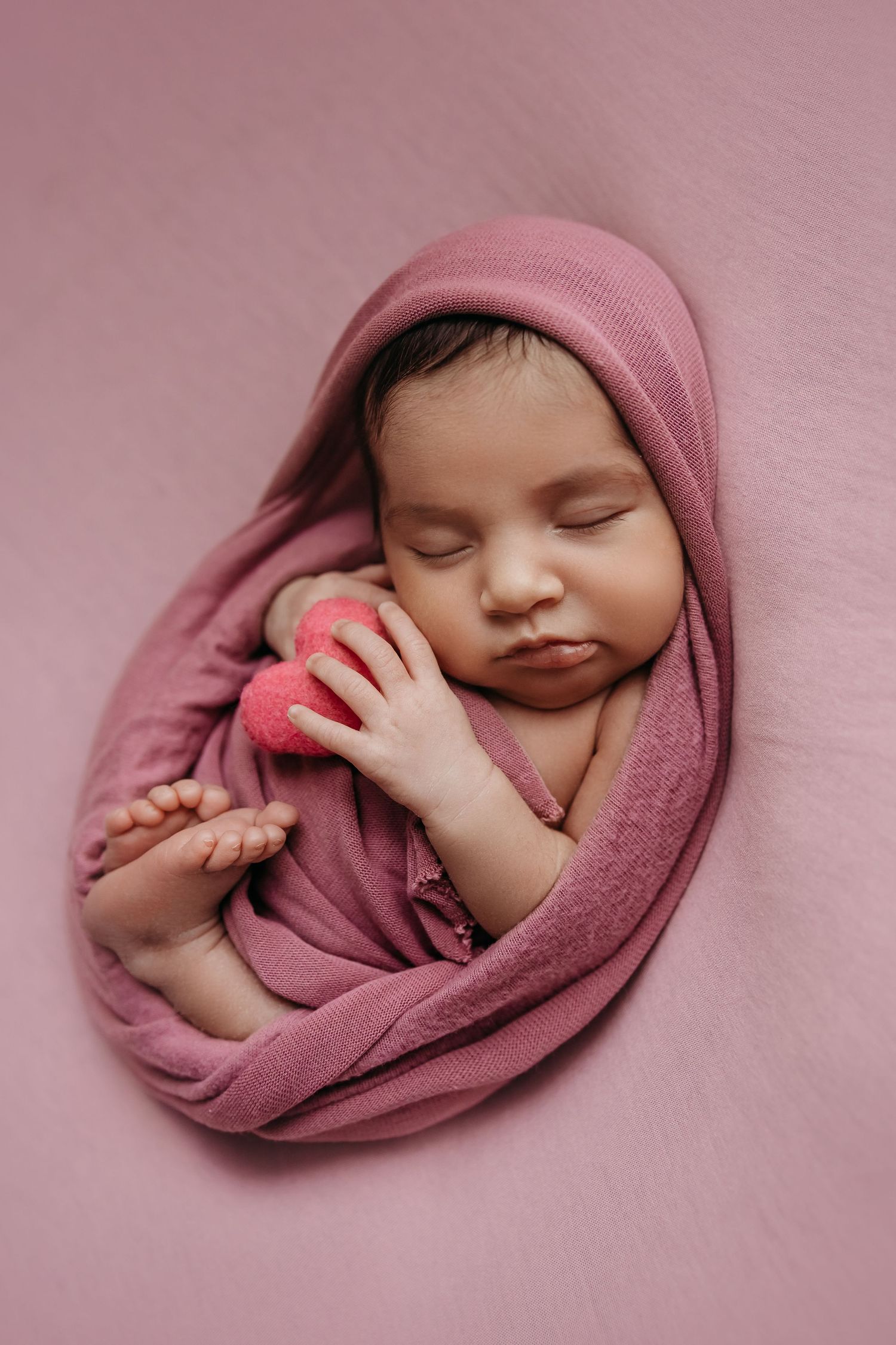 Sleeping newborn wrapped in pink blanket holding small heart prop against pink background.