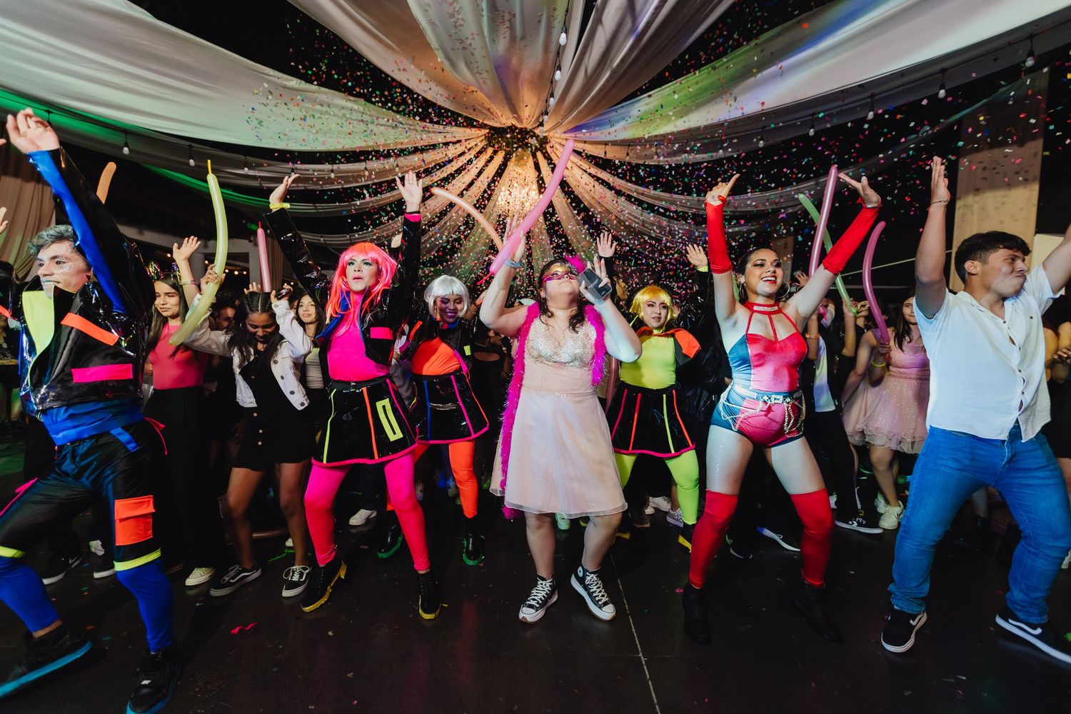 Party guests in colorful costumes dance and celebrate under draped fabric decorations at a festive event.