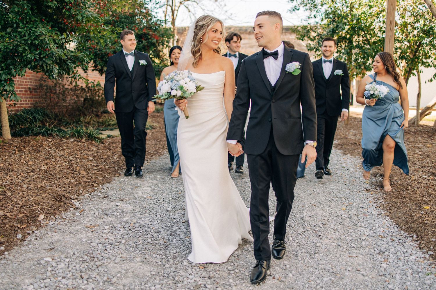 A happy couple walking down a gravel path surrounded by wedding party members in elegant formal attire.