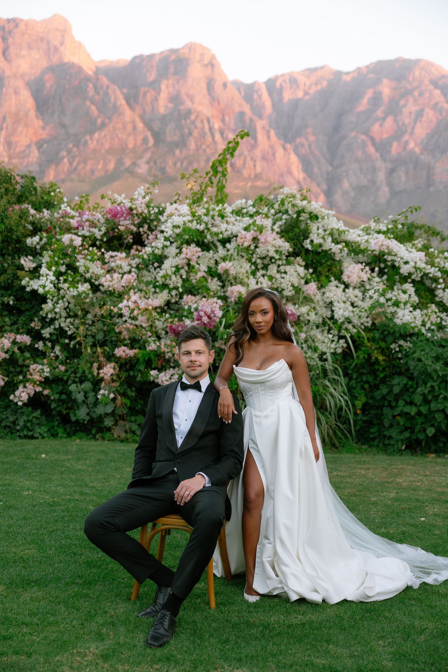 Wedding couple poses in garden setting with blooming white flowers and dramatic mountain backdrop at sunset.