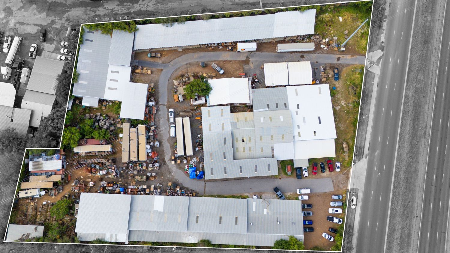 Aerial view of commercial buildings with white roofs and storage yard showing vehicles and equipment on paved lot.