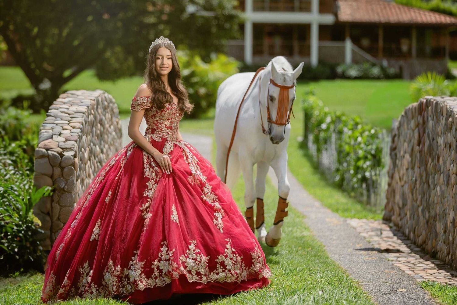 A person in a red and gold embellished formal dress poses along a garden path in a scenic outdoor setting.