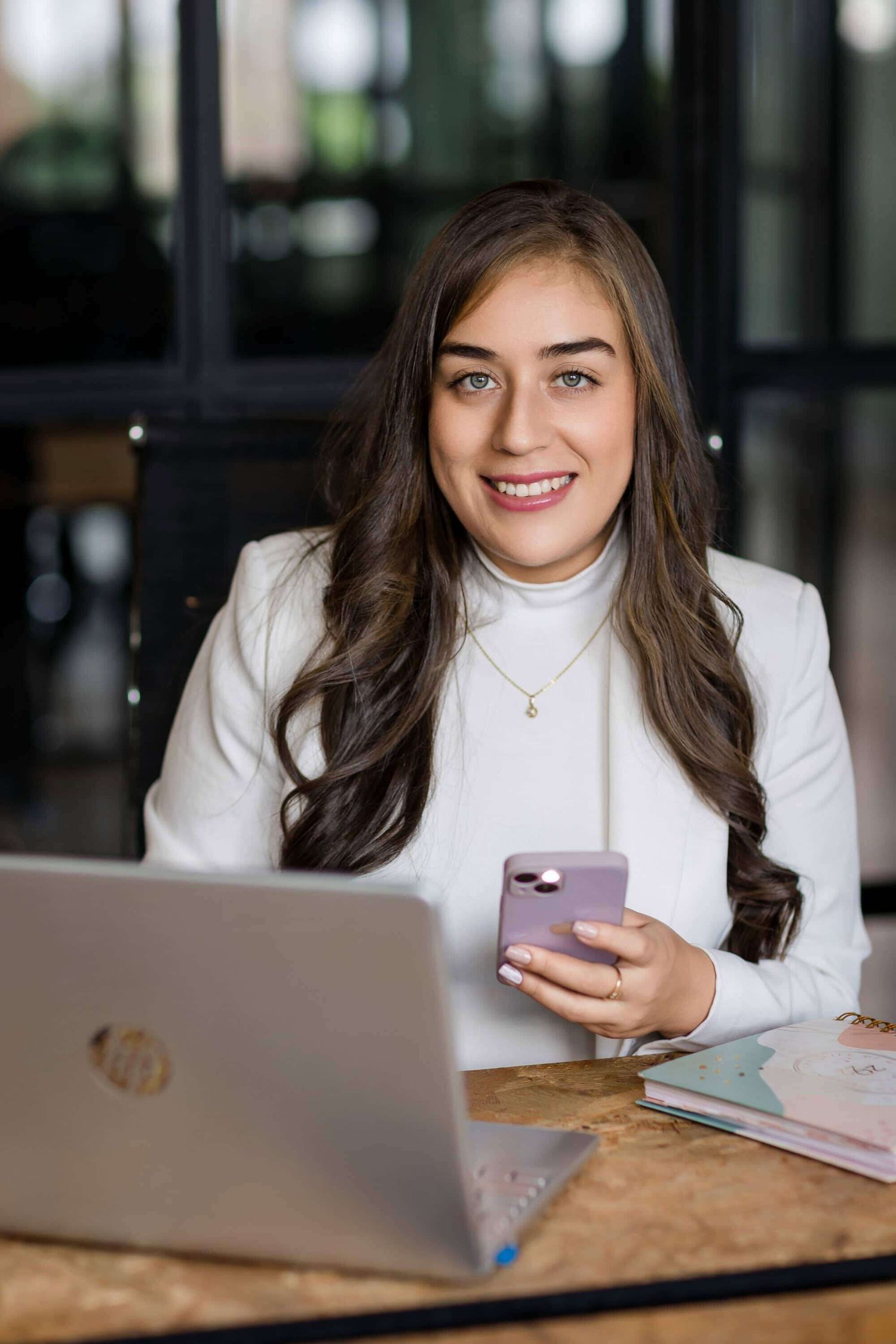 Professional looking person in a white turtleneck sweater working on laptop and checking phone at wooden desk.