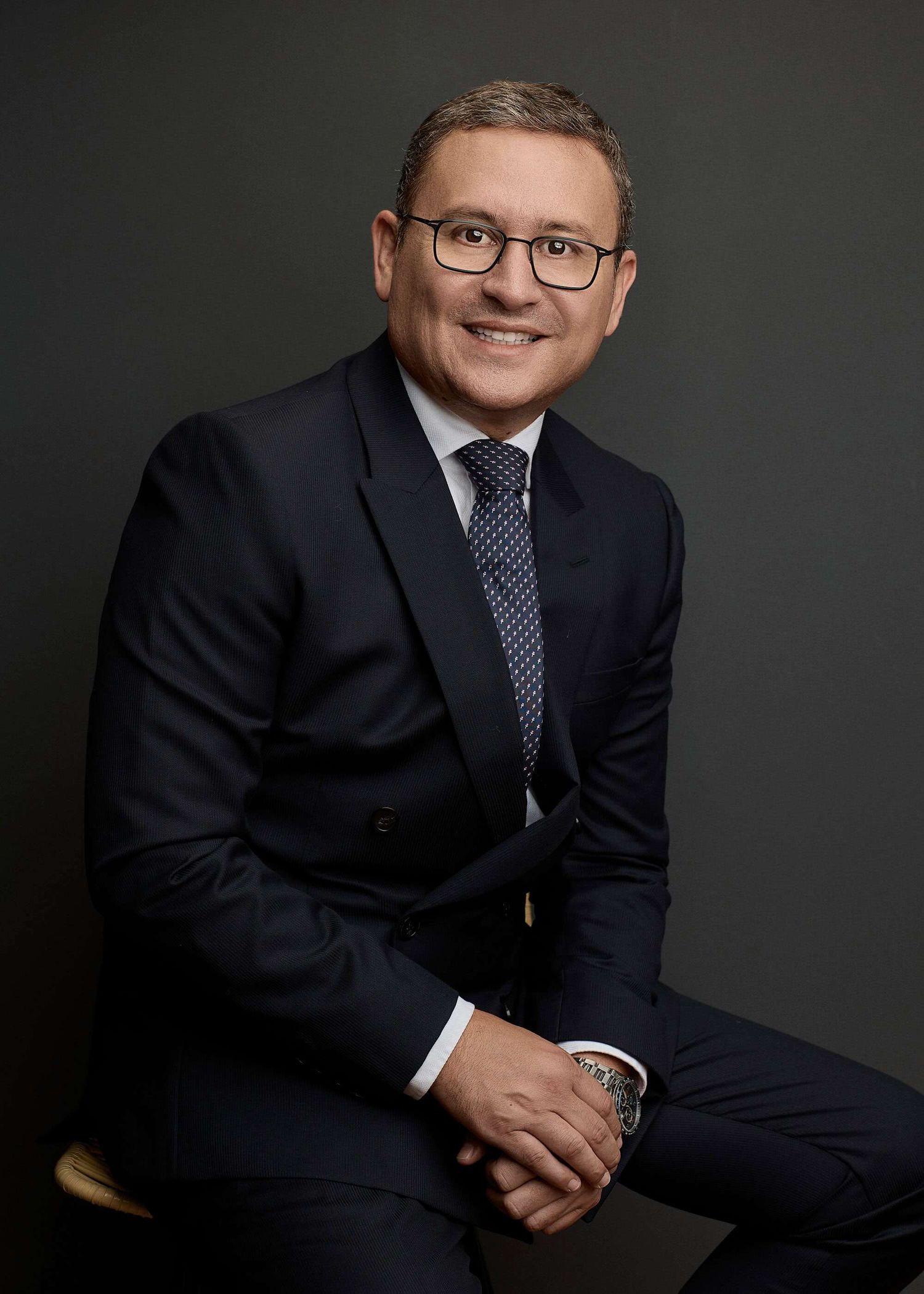 Professional corporate headshot of a person in a dark suit and glasses against a dark gray background.