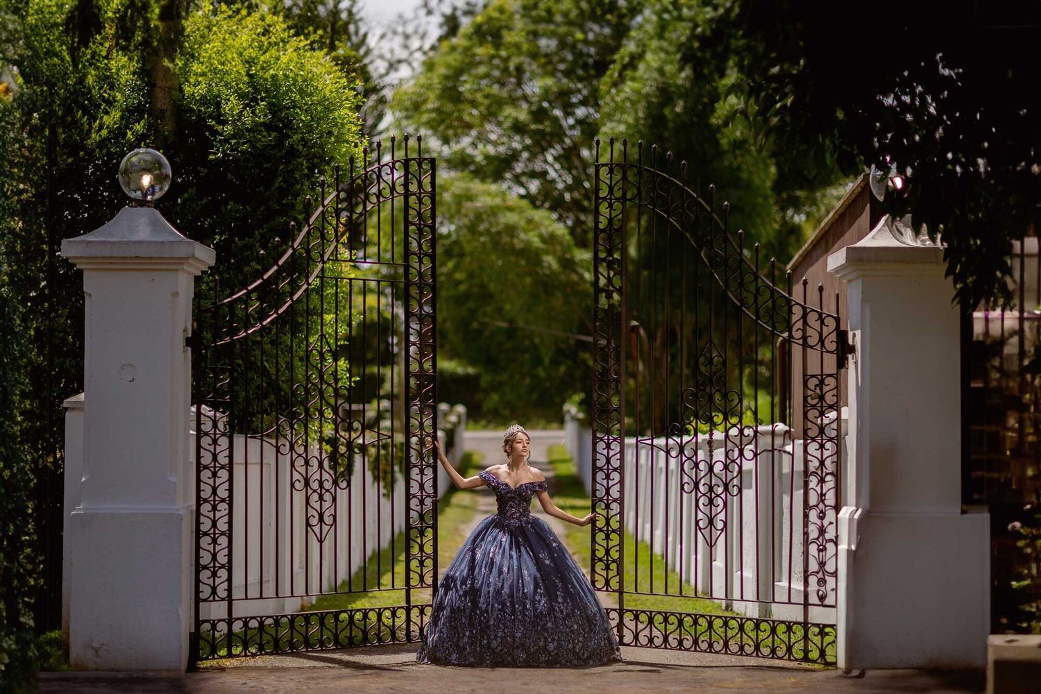 Elegant ball gown seen through ornate white iron gates at a grand estate entrance with lush greenery.