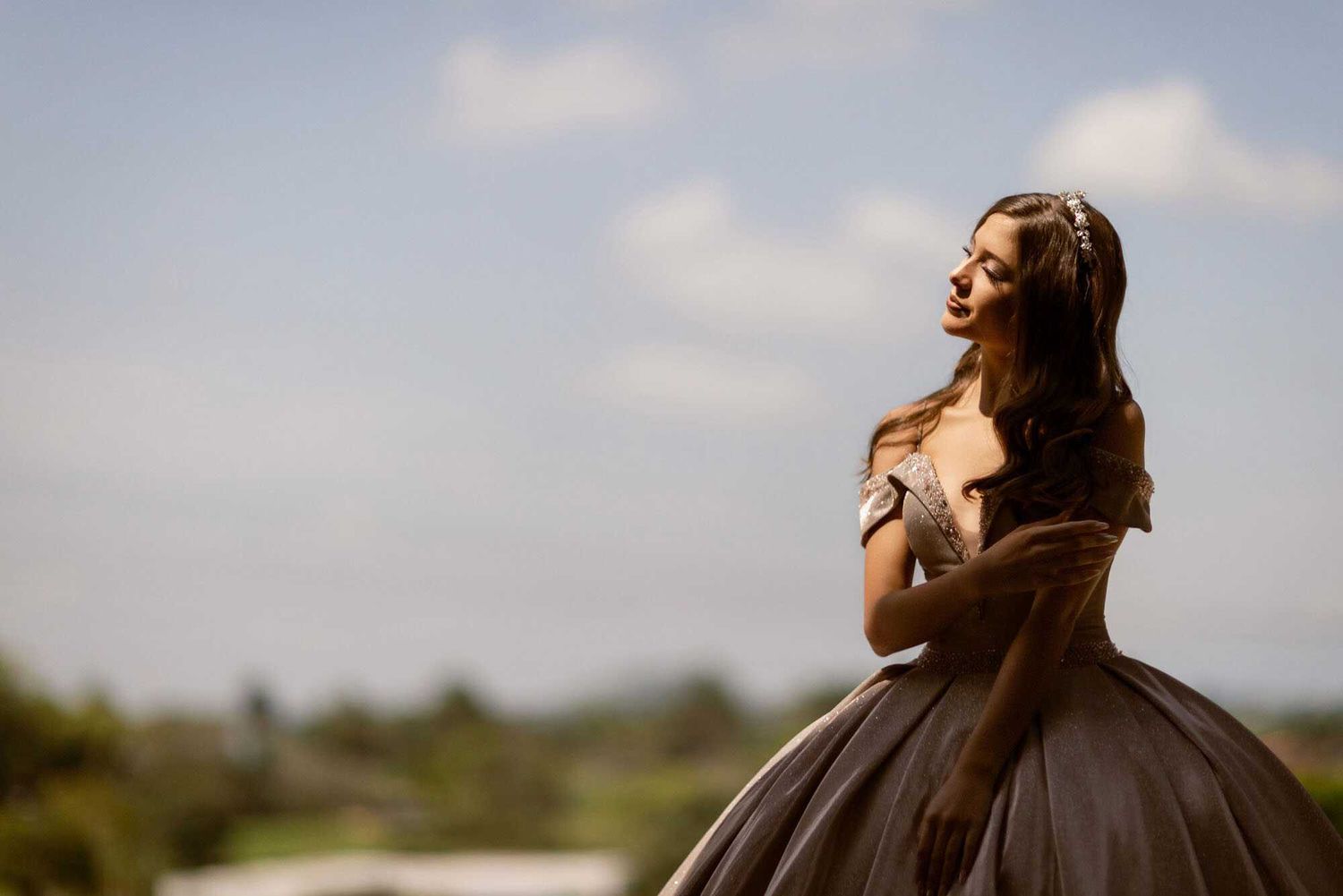 A figure in an elegant brown ballgown poses against a blue sky backdrop with afternoon sunlight.