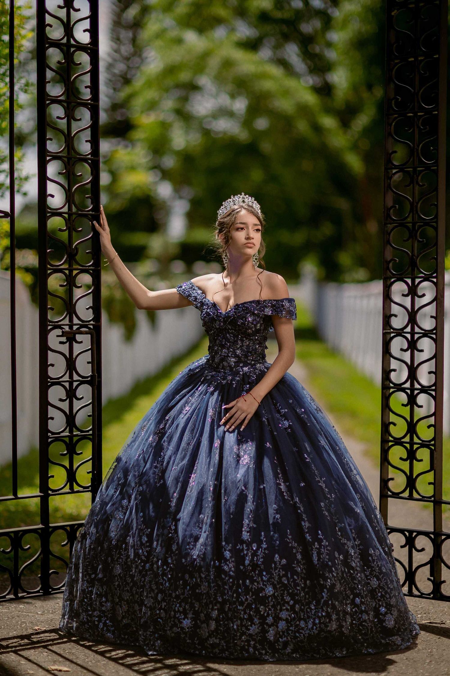 Elegant ball gown in midnight blue sparkles at ornate garden gate with tiara and off-shoulder neckline catching evening light.