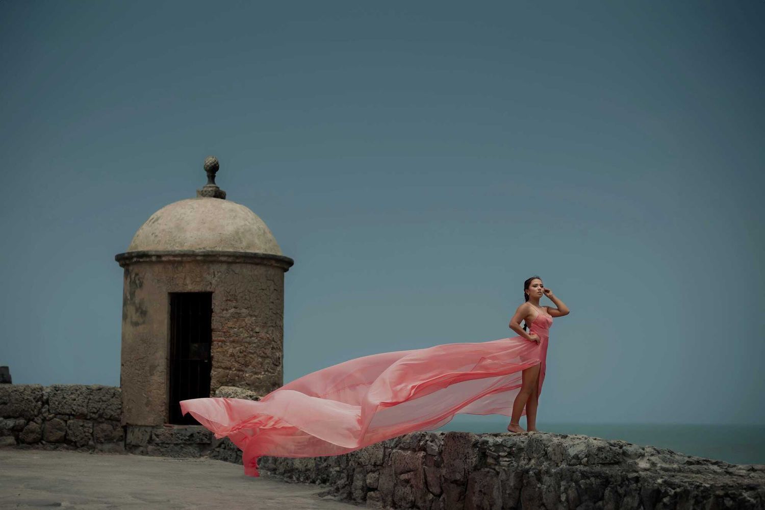 Person in flowing pink dress poses on historic stone fortress wall with domed watchtower against blue sky and ocean backdrop.