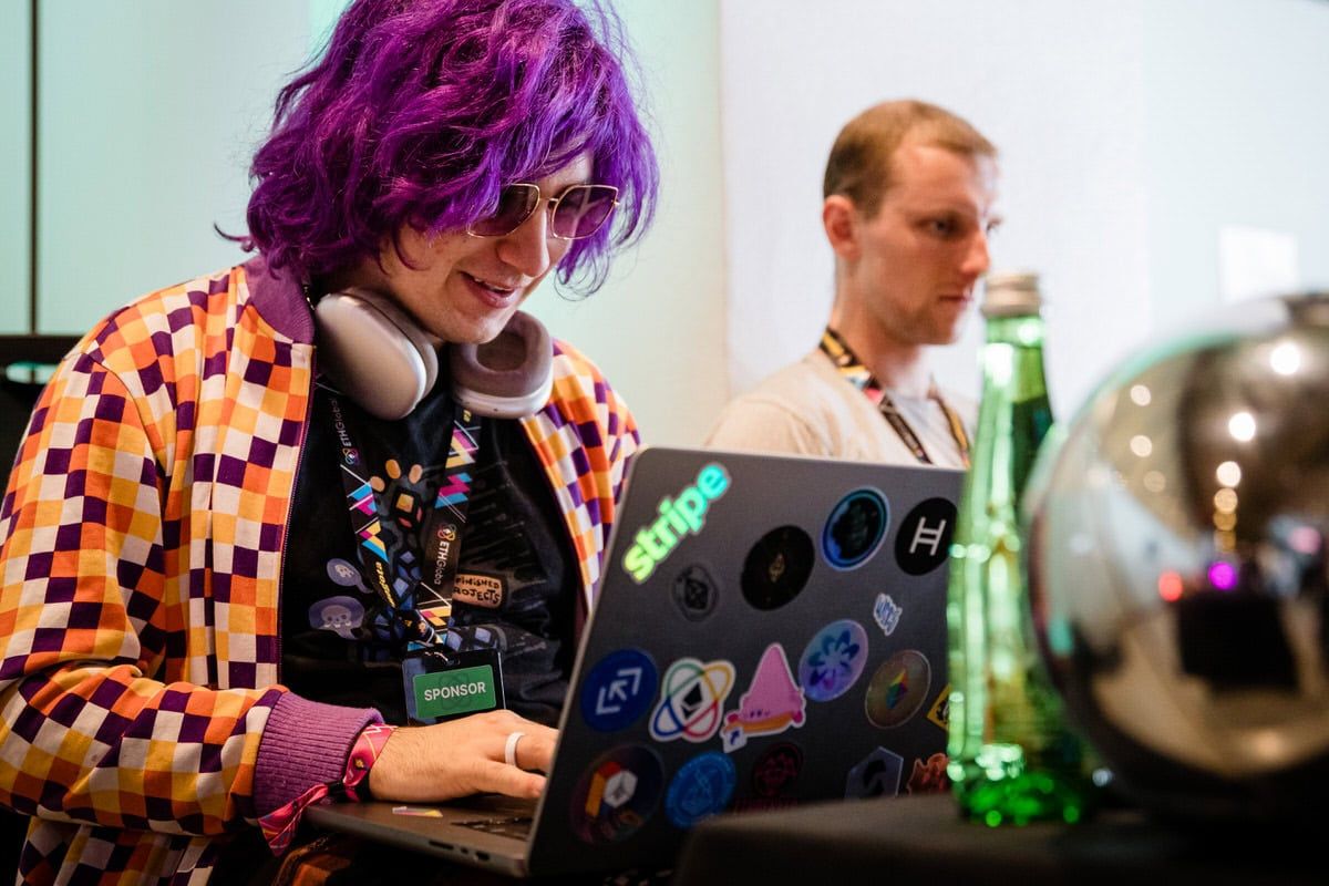 Person with vibrant purple hair and checkered jacket uses laptop covered in colorful stickers while wearing headphones.