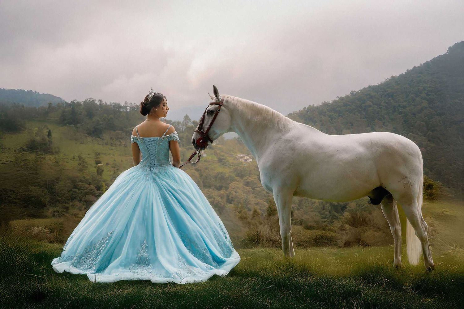 A bride in a flowing blue dress poses with a majestic white horse against scenic mountain landscapes at sunset.