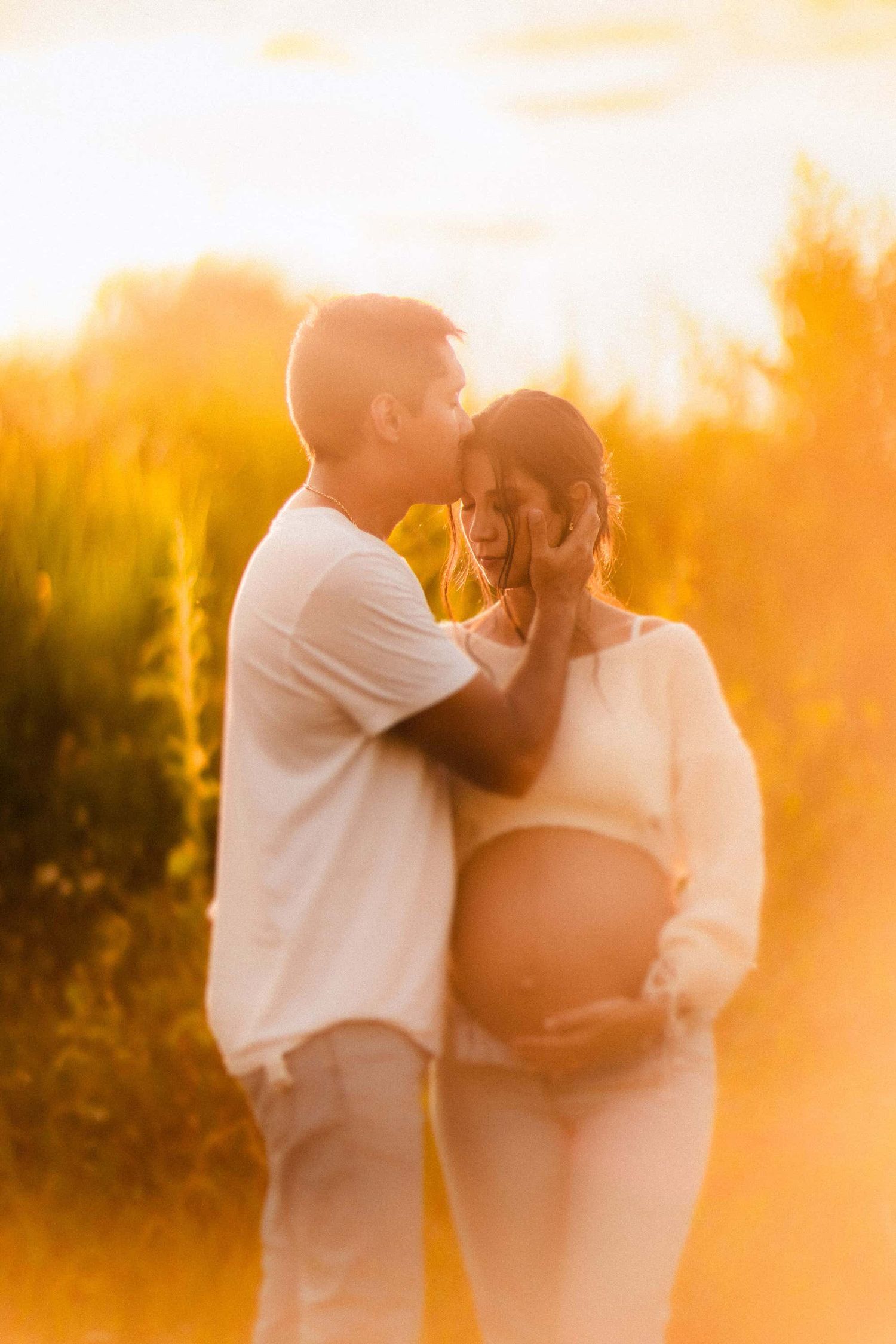 A couple shares an intimate moment during a maternity photoshoot at sunset with warm golden light in the background.