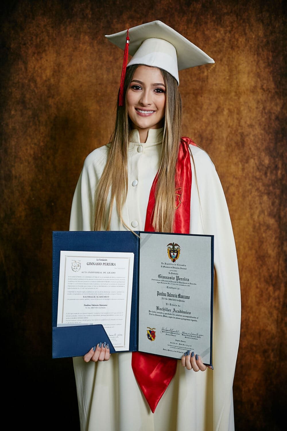 Graduado con gorra blanca y vestido con roble rojo sostiene dos diplomas contra el telón de fondo marrón. Graduada mostrando diploma