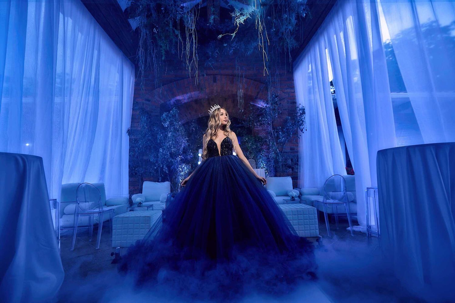 A figure in a dramatic navy blue ball gown stands in an ethereal hallway with flowing white curtains and magical lighting.