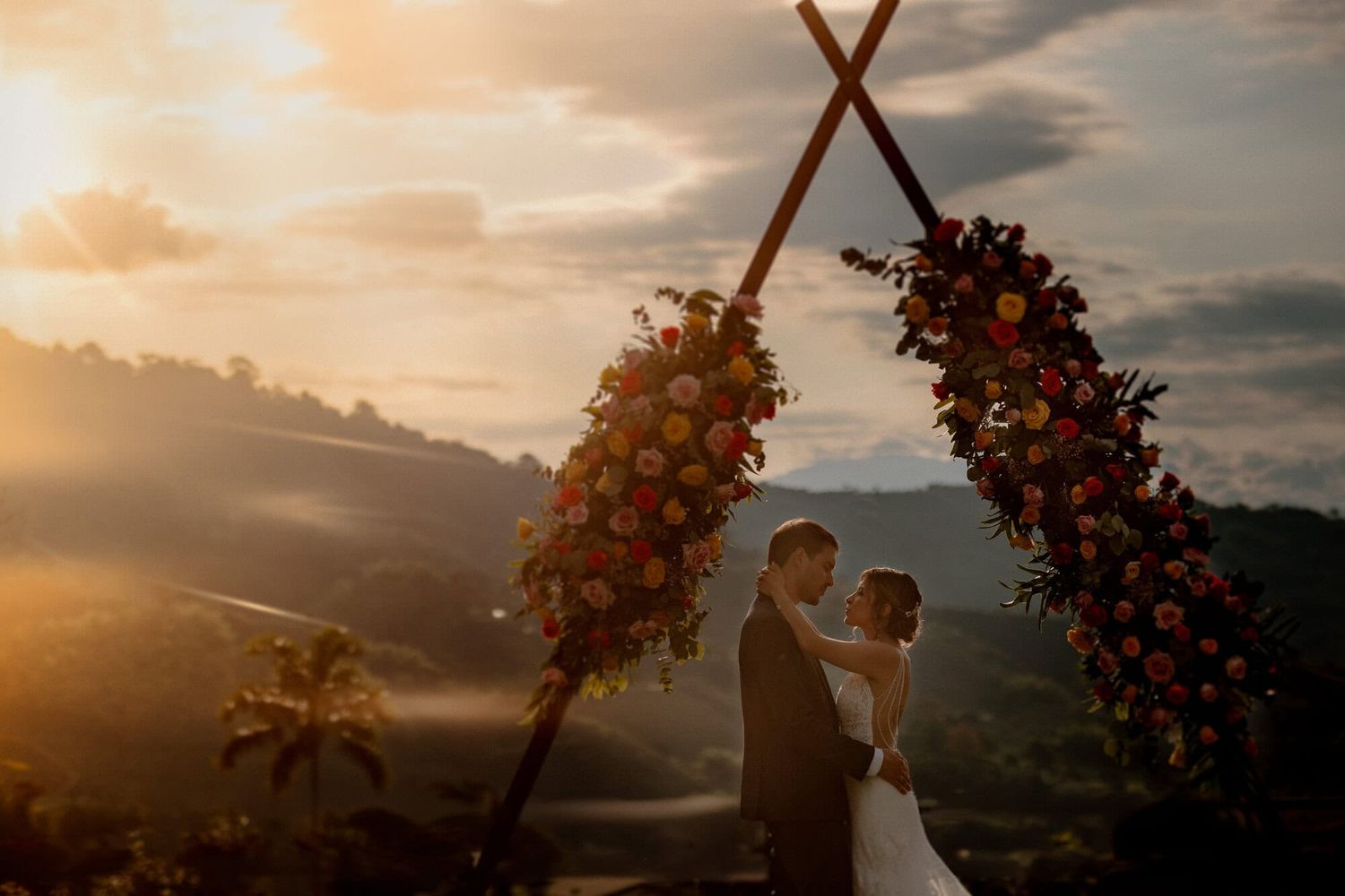 A romantic sunset wedding ceremony under a rustic wooden arch adorned with red and orange flowers in the mountains.