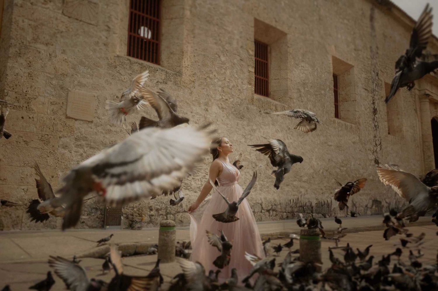 A person in a flowing pink dress walks through a flock of pigeons taking flight in front of an old stone building.