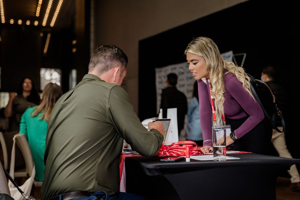 Two people at a table during what appears to be a book signing event with a red book display.