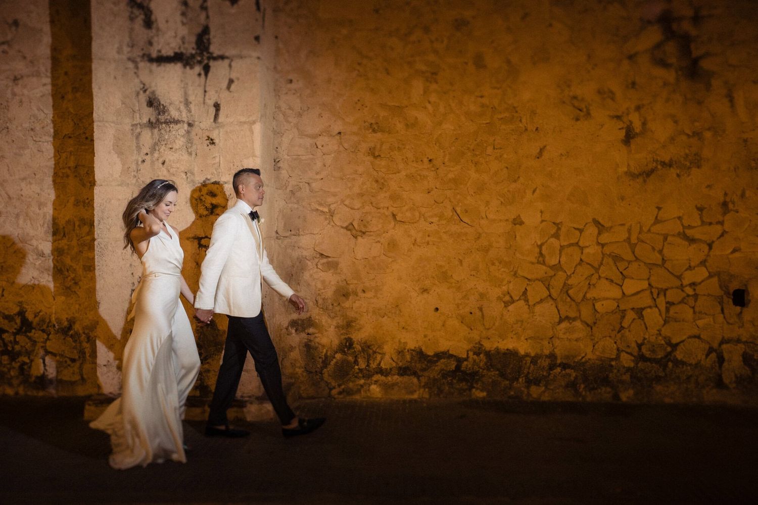 A couple in wedding attire walks along a rustic orange wall at dusk, creating a romantic silhouette effect.
