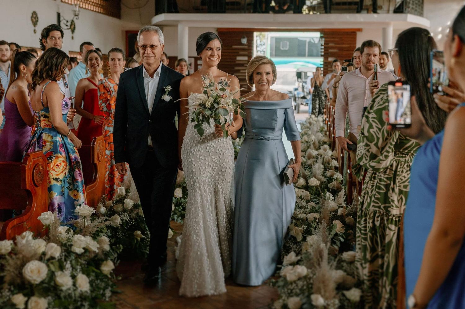 Novia acompañada de sus padres entrando a la iglesia, rodeada de flores y emociones.