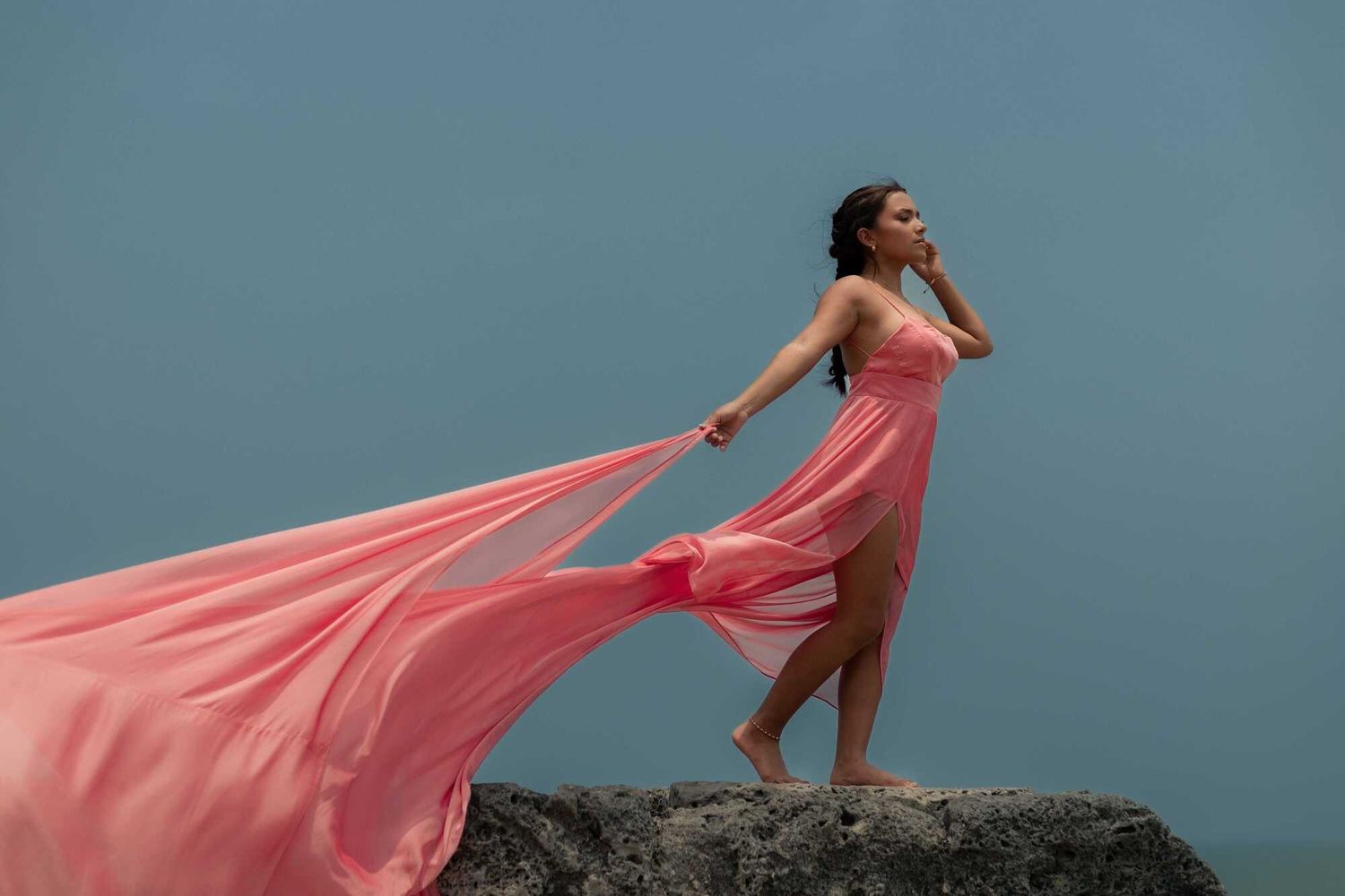 Person in flowing coral dress poses dramatically on coastal rocks against moody blue sky at sunset.