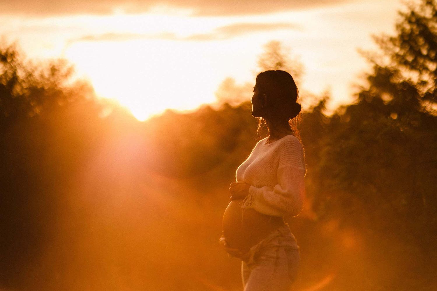 A silhouette of a pregnant woman standing in golden sunset light with trees in the background.