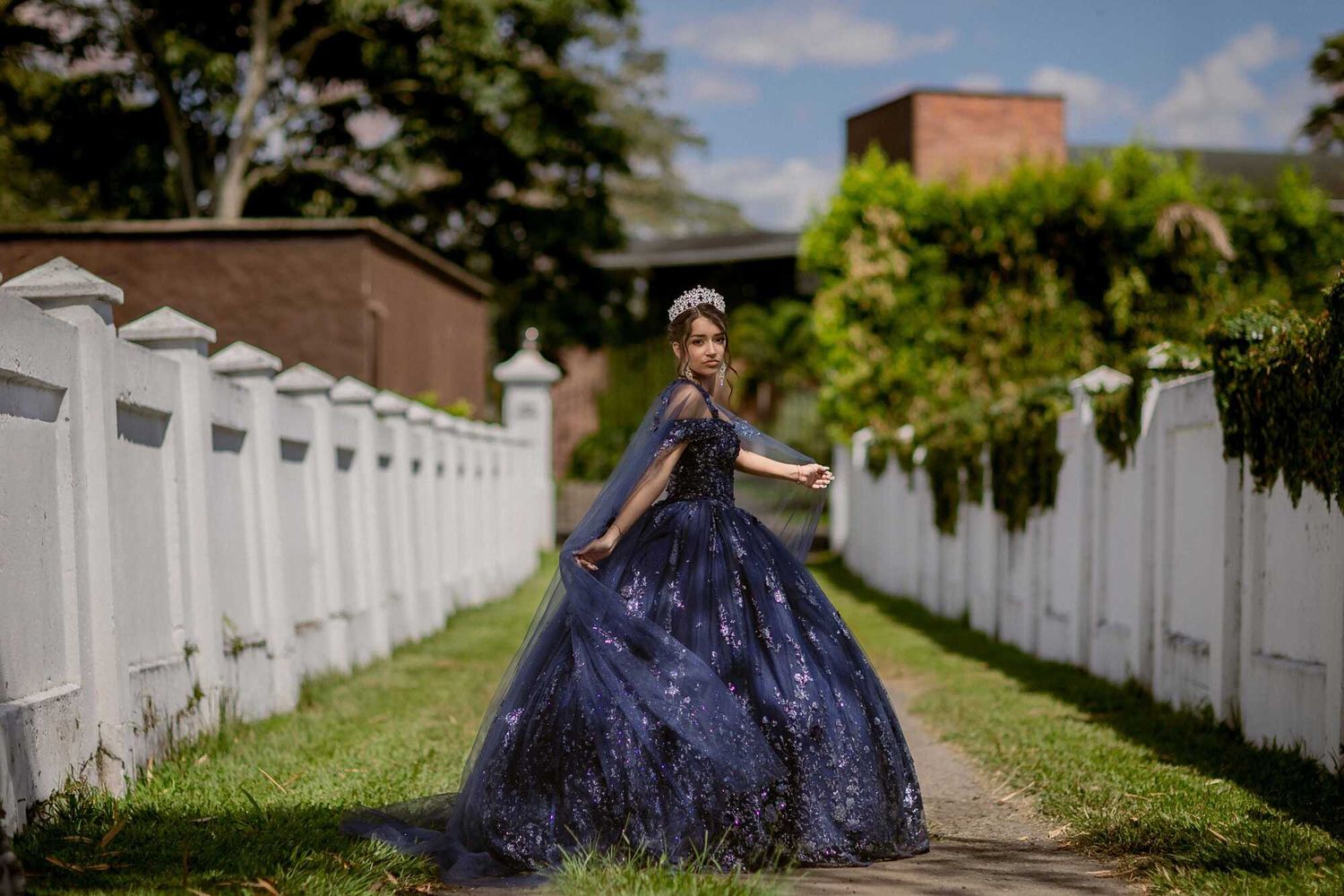 Person in a flowing navy blue gown walks along a classical white fenced garden walkway.