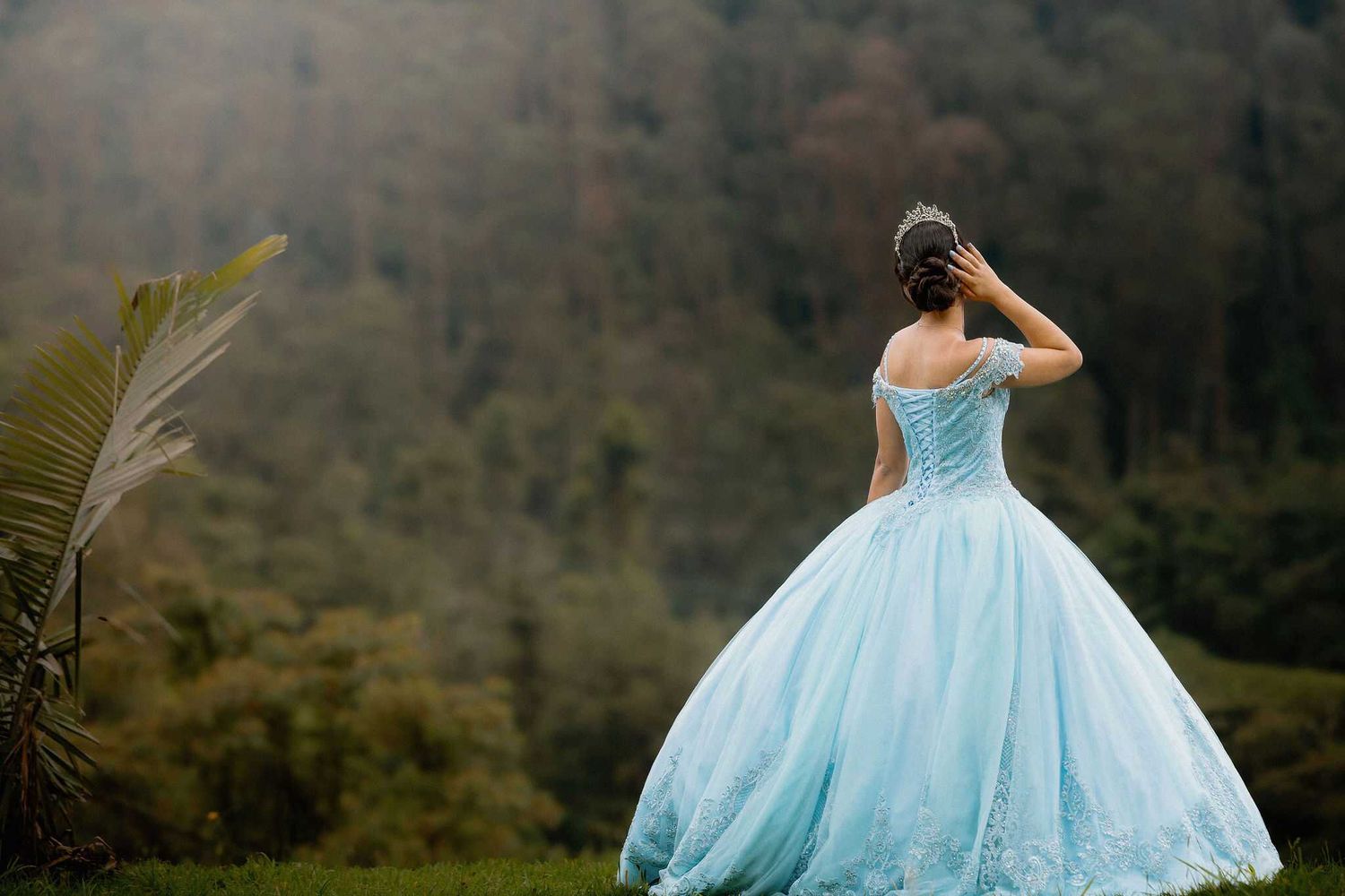 A person in a light blue ball gown stands looking out at misty mountains in a dreamy outdoor setting.