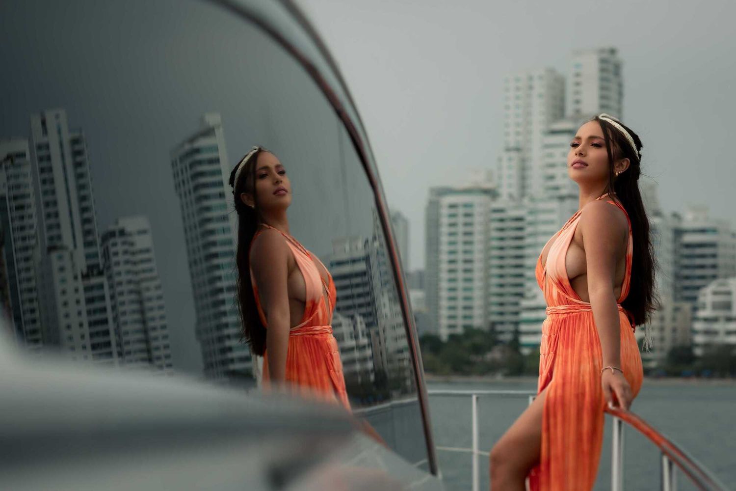 Woman in coral dress poses against urban skyline backdrop reflected in car mirror.