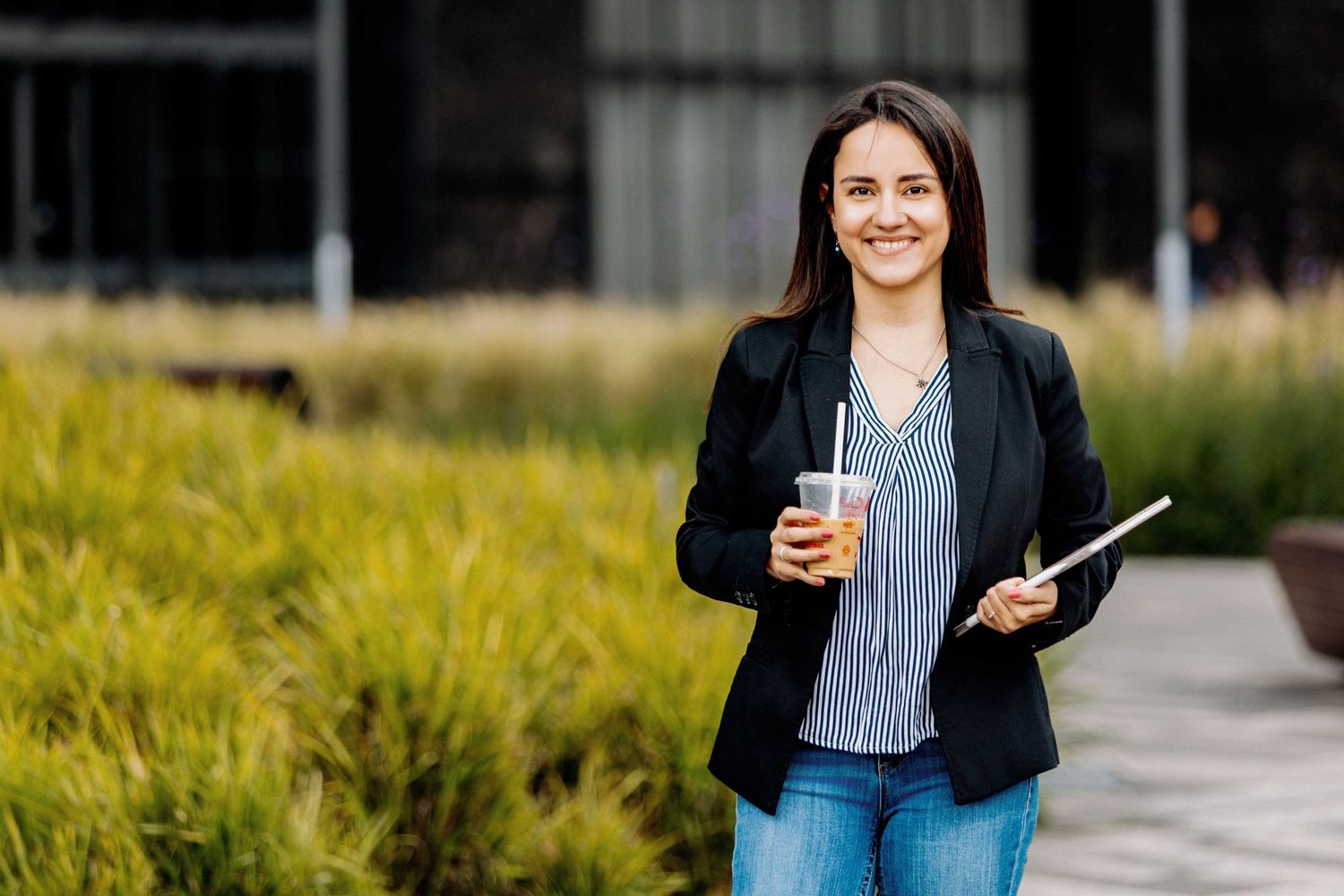 Young professional in a black blazer and jeans holds a coffee and tablet while standing outdoors near landscaping.