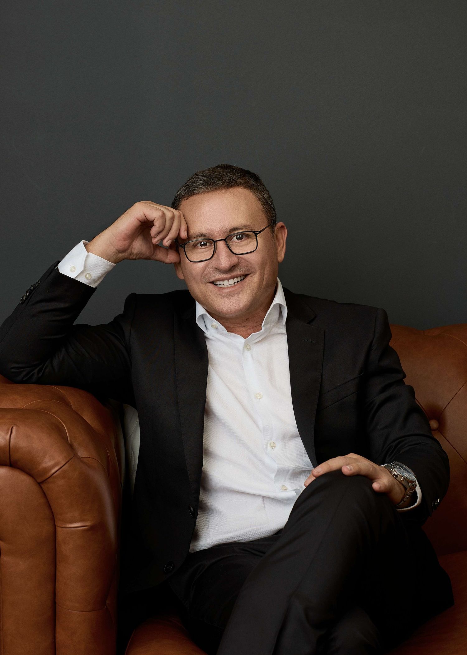 Professional studio portrait of someone in a dark suit and white shirt sitting casually on a brown leather chair against gray backdrop.